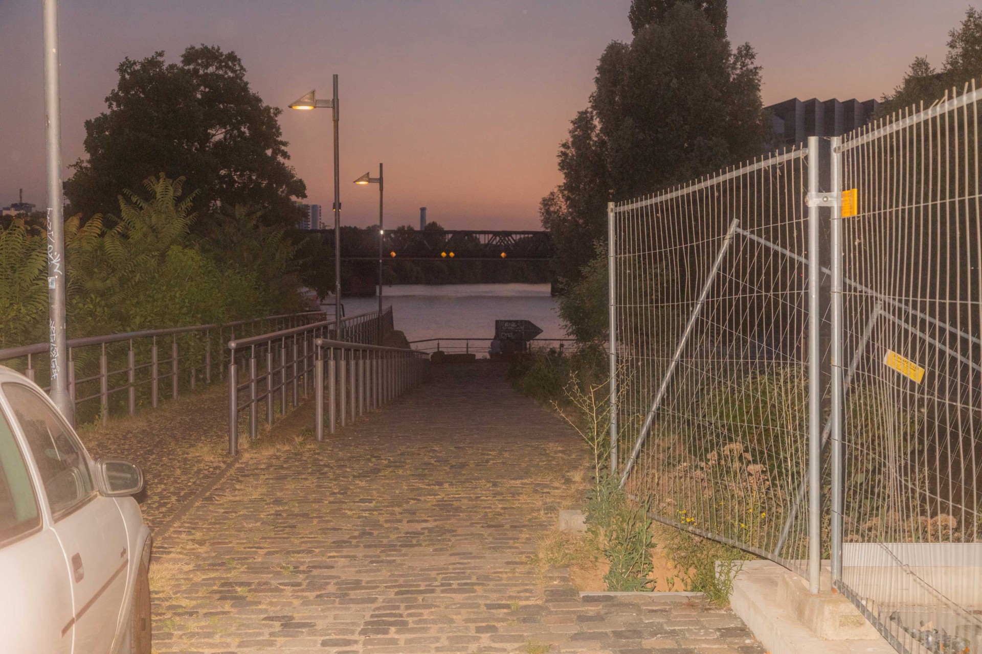 Kopfsteinpflasterpfade führen zum Wasser, beleuchtet von Straßenlaternen, umgeben von Bäumen und einem Zaun im Abendlicht.