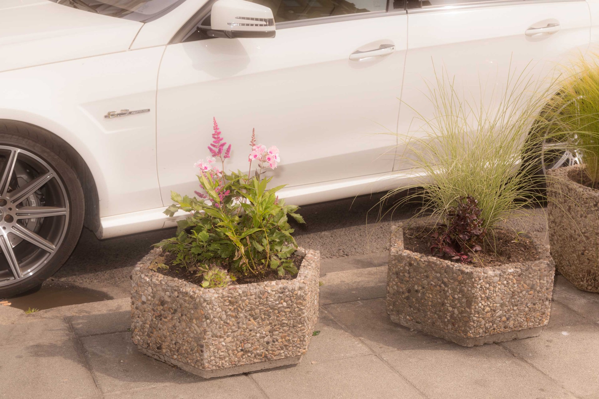 Weißes Auto neben blühenden Pflanzen in hexagonalen Betontöpfen auf einer gepflasterten Straße.