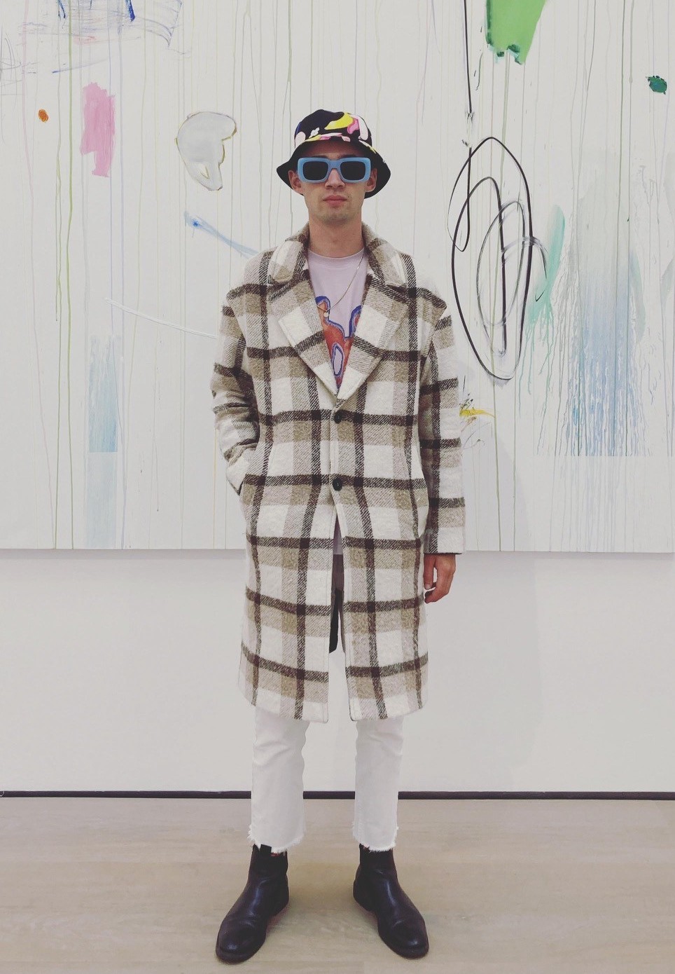 Junger Mann im gestreiften Mantel und Sonnenbrille vor einem bunten Kunstwerk in einer Galerie.