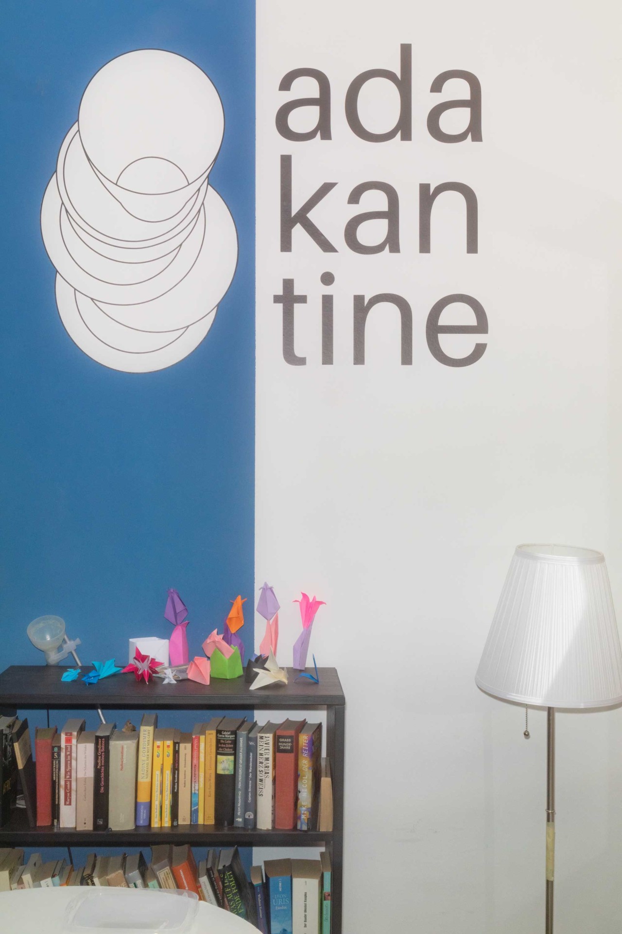 Moderne Wandgestaltung mit dem Schriftzug "ada kantine", gestapelten Tellern und bunten Origami-Figuren auf einem Regal.