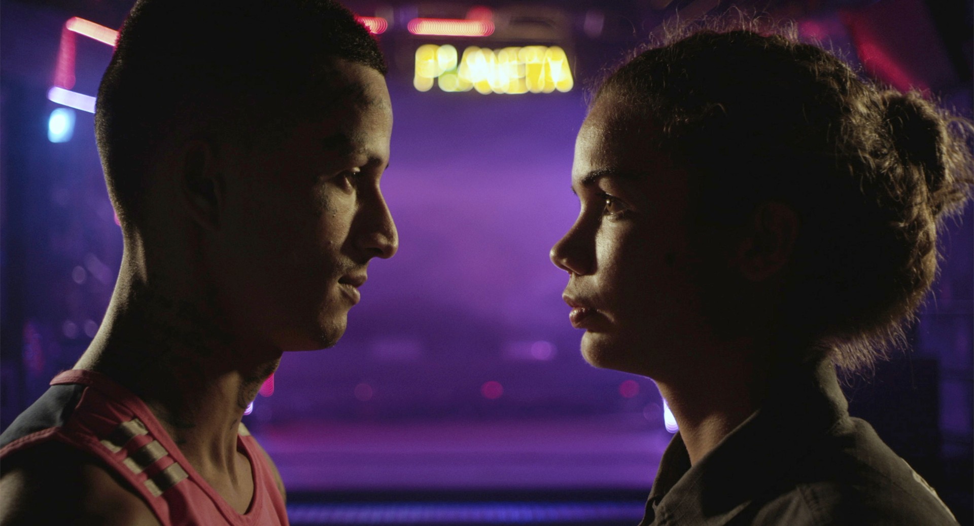 Zwei Personen stehen sich gegenüber, blicken sich intensiv an, vor einem bunten, unscharfen Hintergrund.