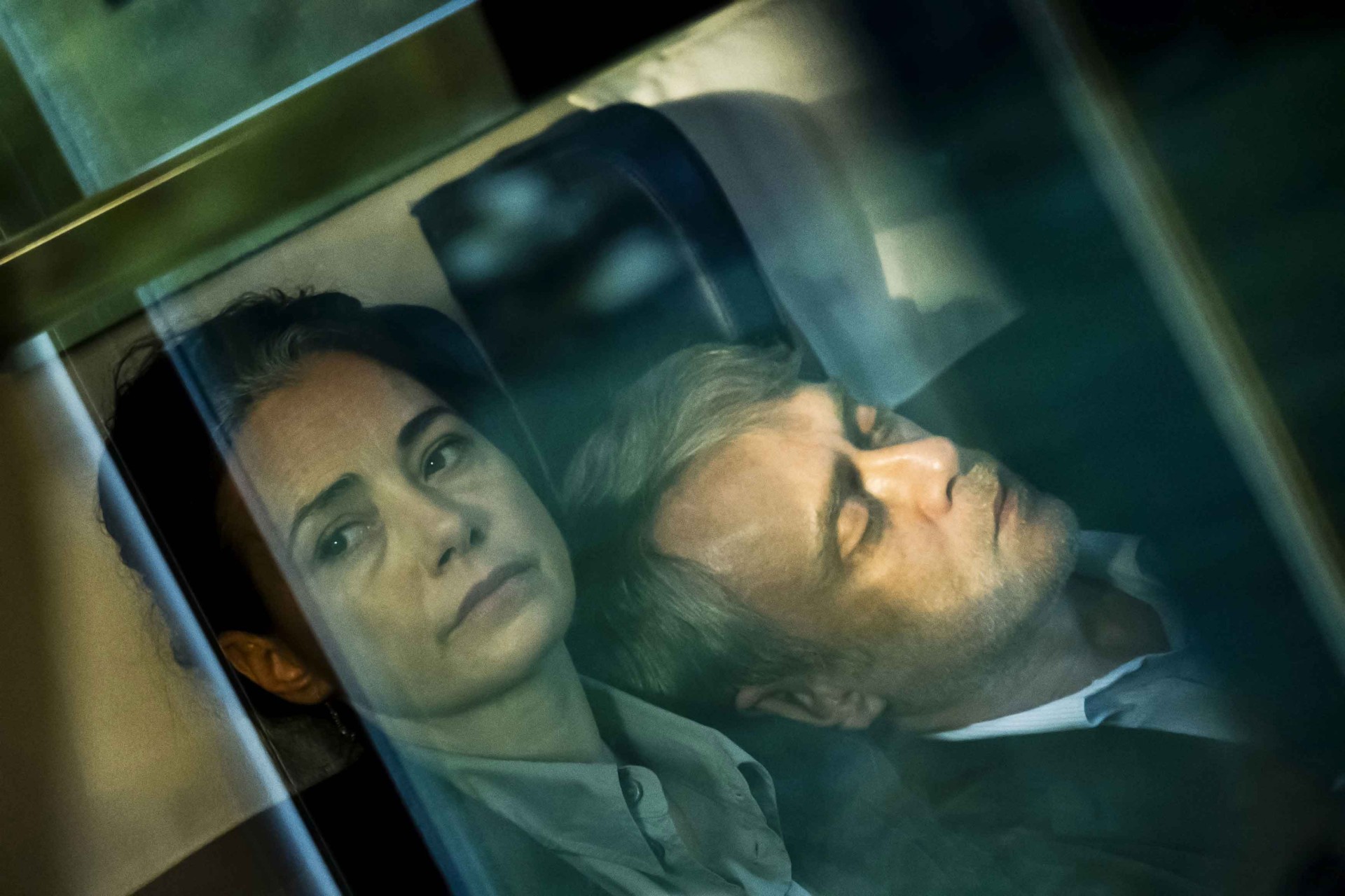 Eine nachdenkliche Frau sitzt neben einem schlafenden Mann in einem Fahrzeug, während im Hintergrund die Fenster reflektieren.