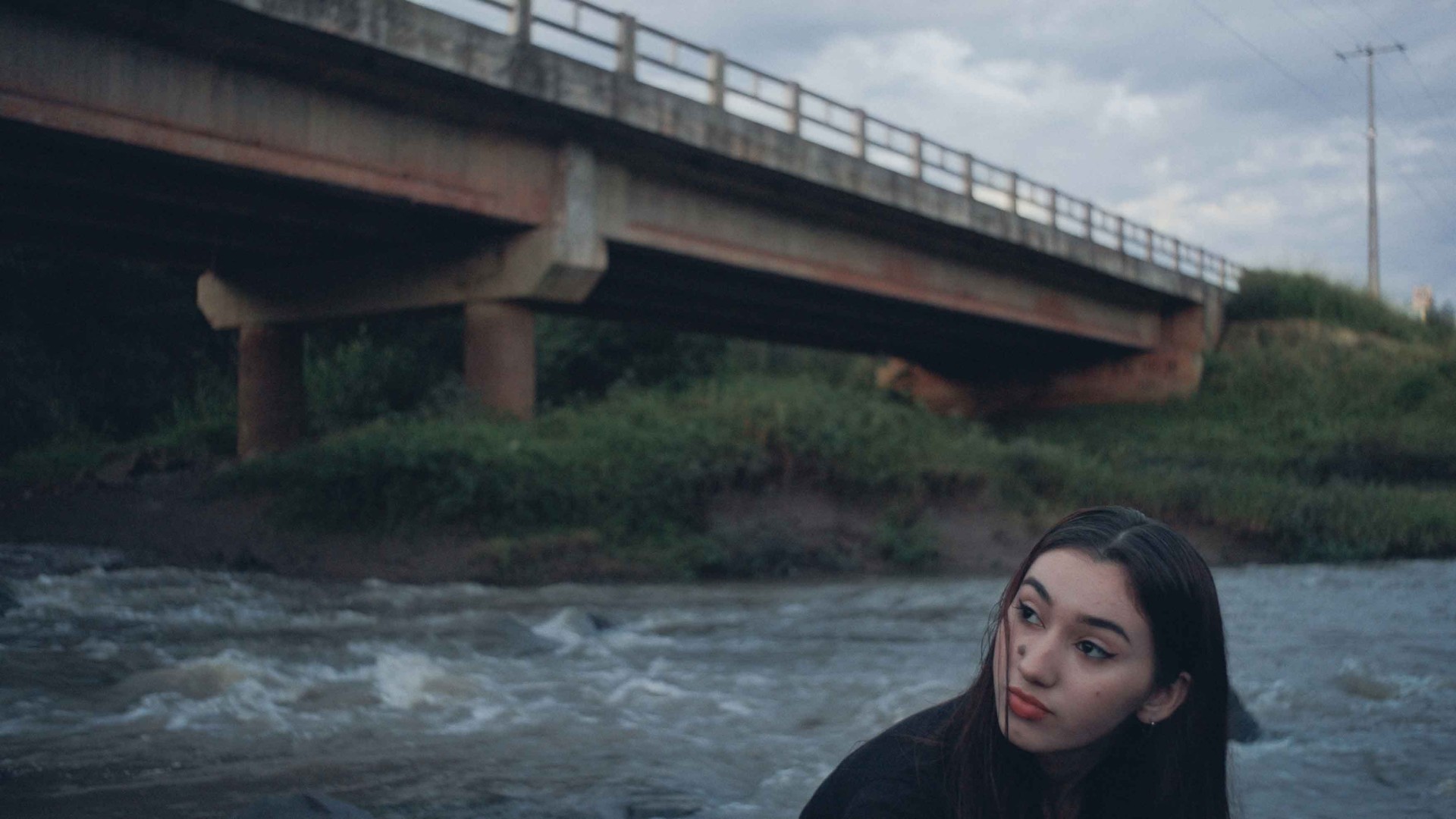 Eine junge Frau sitzt am Ufer unter einer Brücke, im Hintergrund fließt Wasser und der Himmel ist bewölkt.