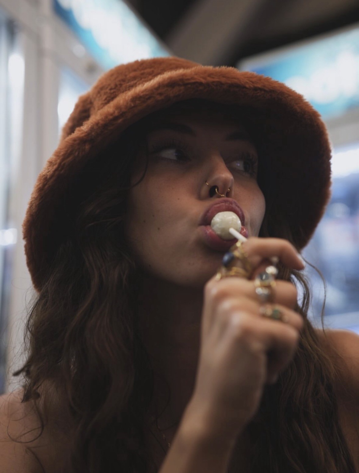 Young woman with curly hair, brown hat, and nose ring, licking an ice cream. Background is blurry.