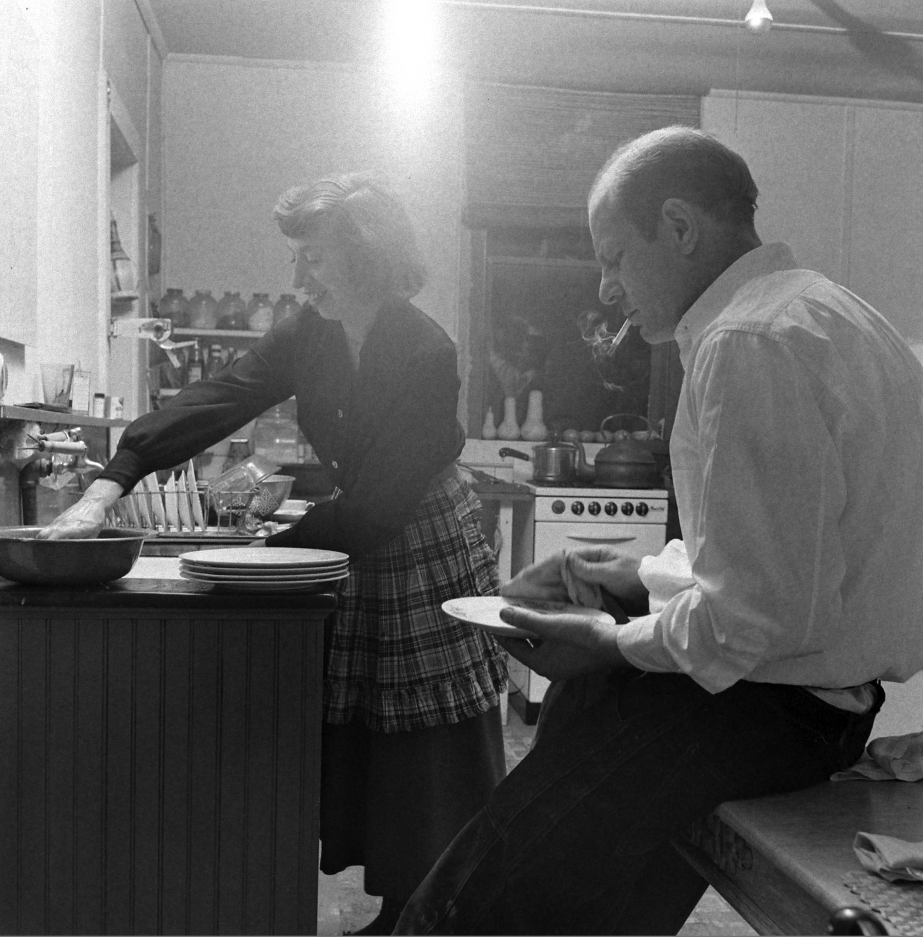 A man sits at a table while a woman washes dishes in the kitchen. Black and white photo.