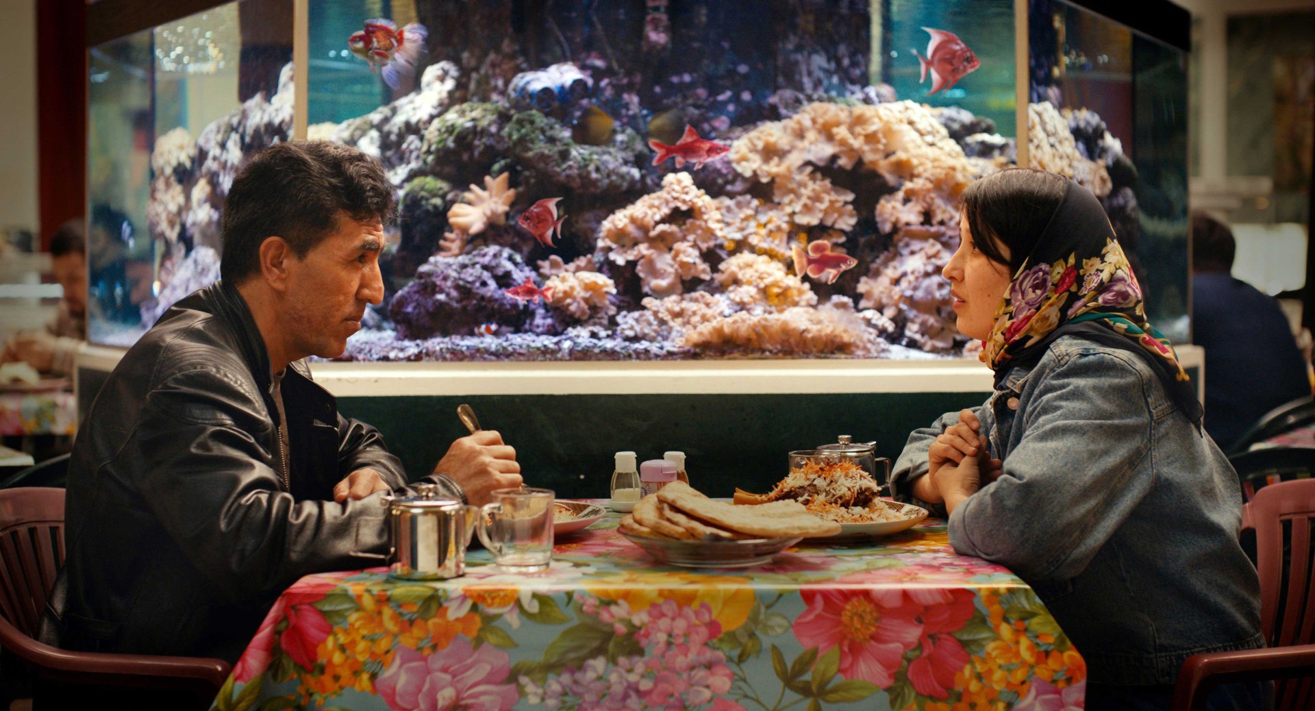 Ein Paar sitzt an einem Tisch mit Essen, während im Hintergrund ein Aquarium mit Fischen zu sehen ist.