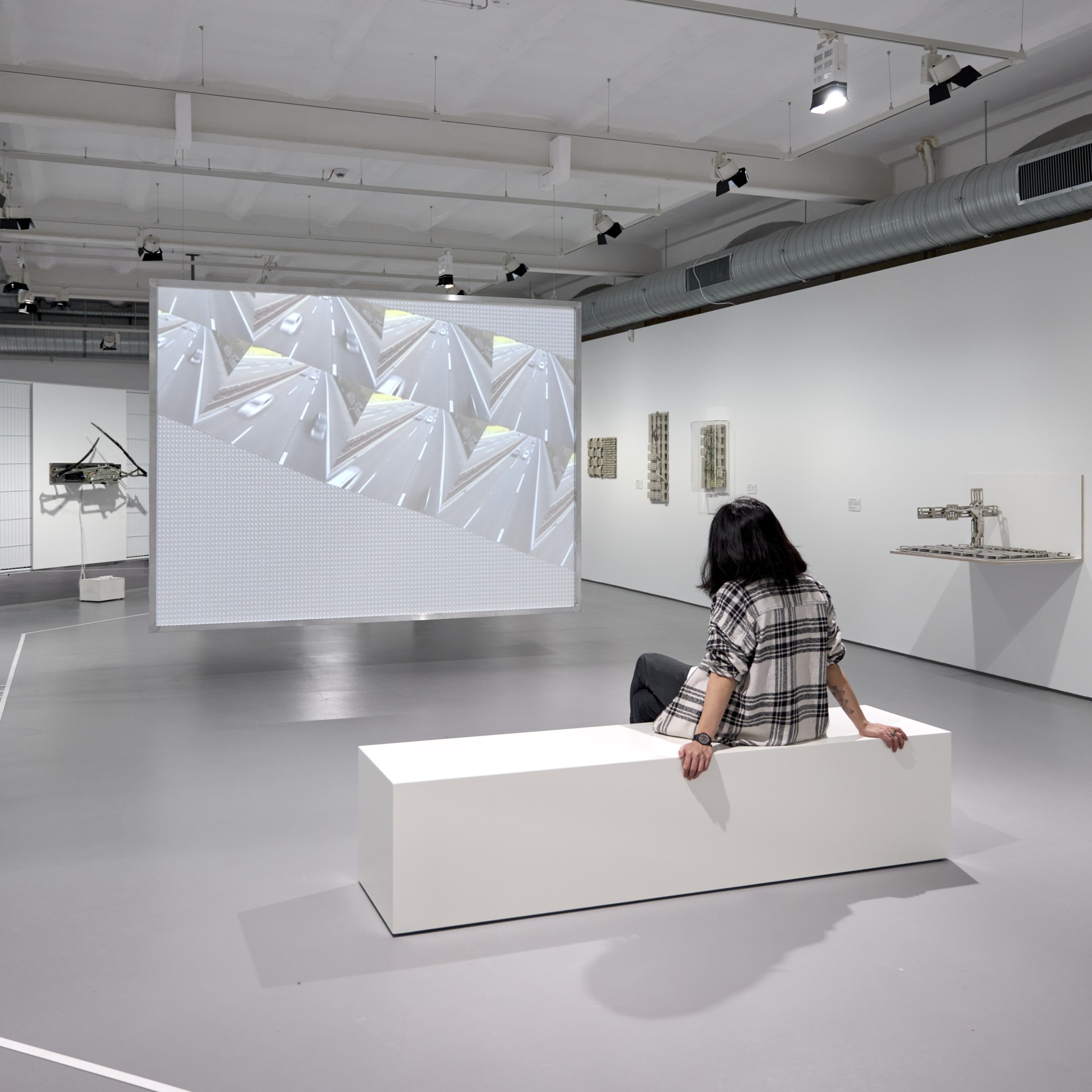 Person sitzt auf einer weißen Bank in einem modernen Ausstellungsraum, während an einer Wand ein Video abgespielt wird.