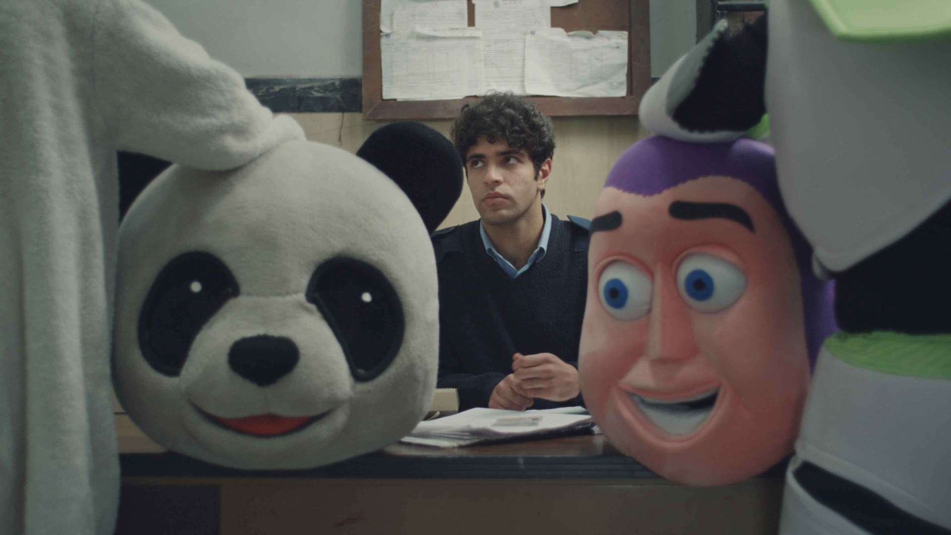 Ein junger Mann sitzt zwischen zwei großen Maskottchenköpfen: einem Panda und einem bunten, fröhlichen Charakter.