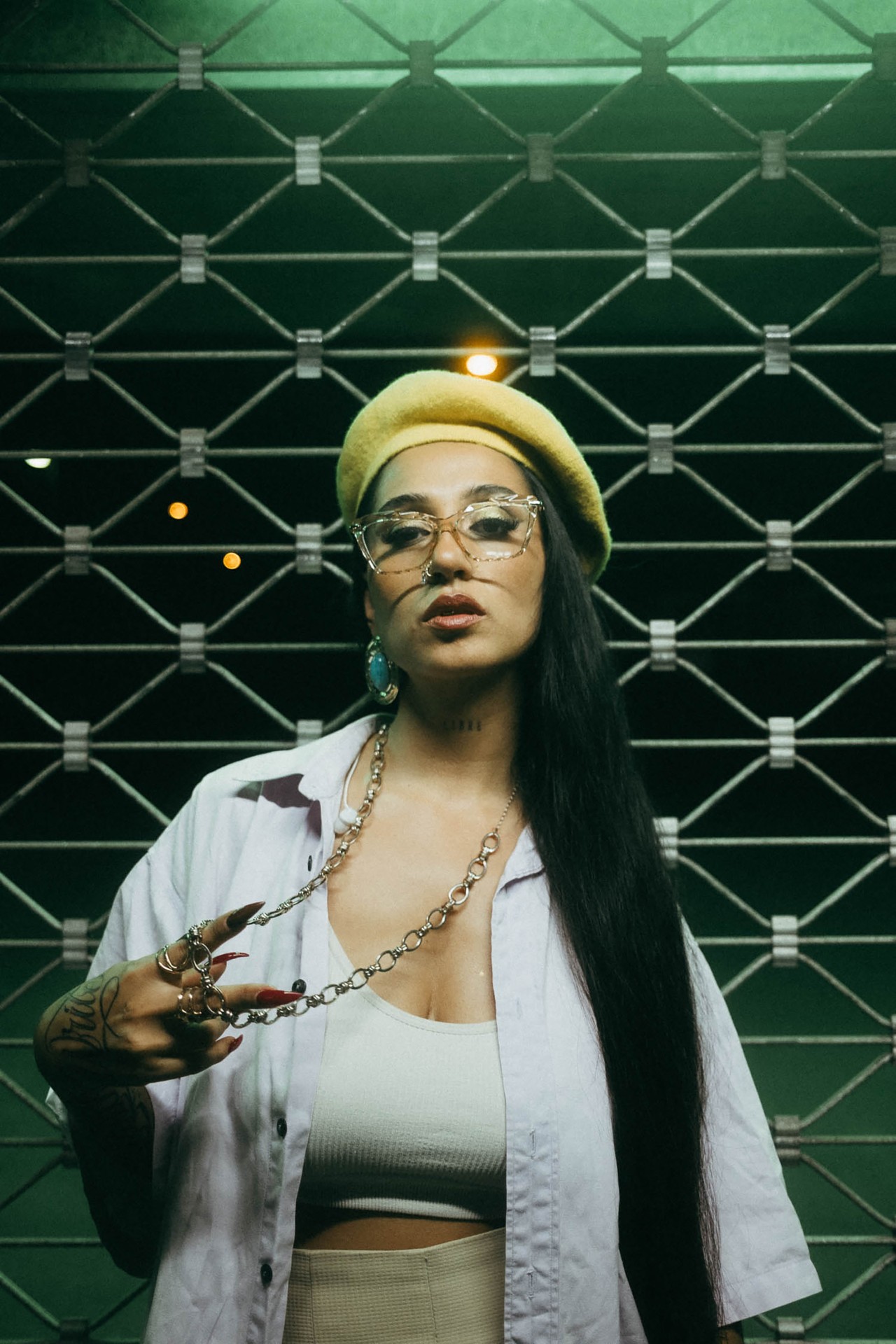 Stylische Person mit langen Haaren, gelbem Hut und Brille, posiert vor einem Gitter mit urbanem Charme.