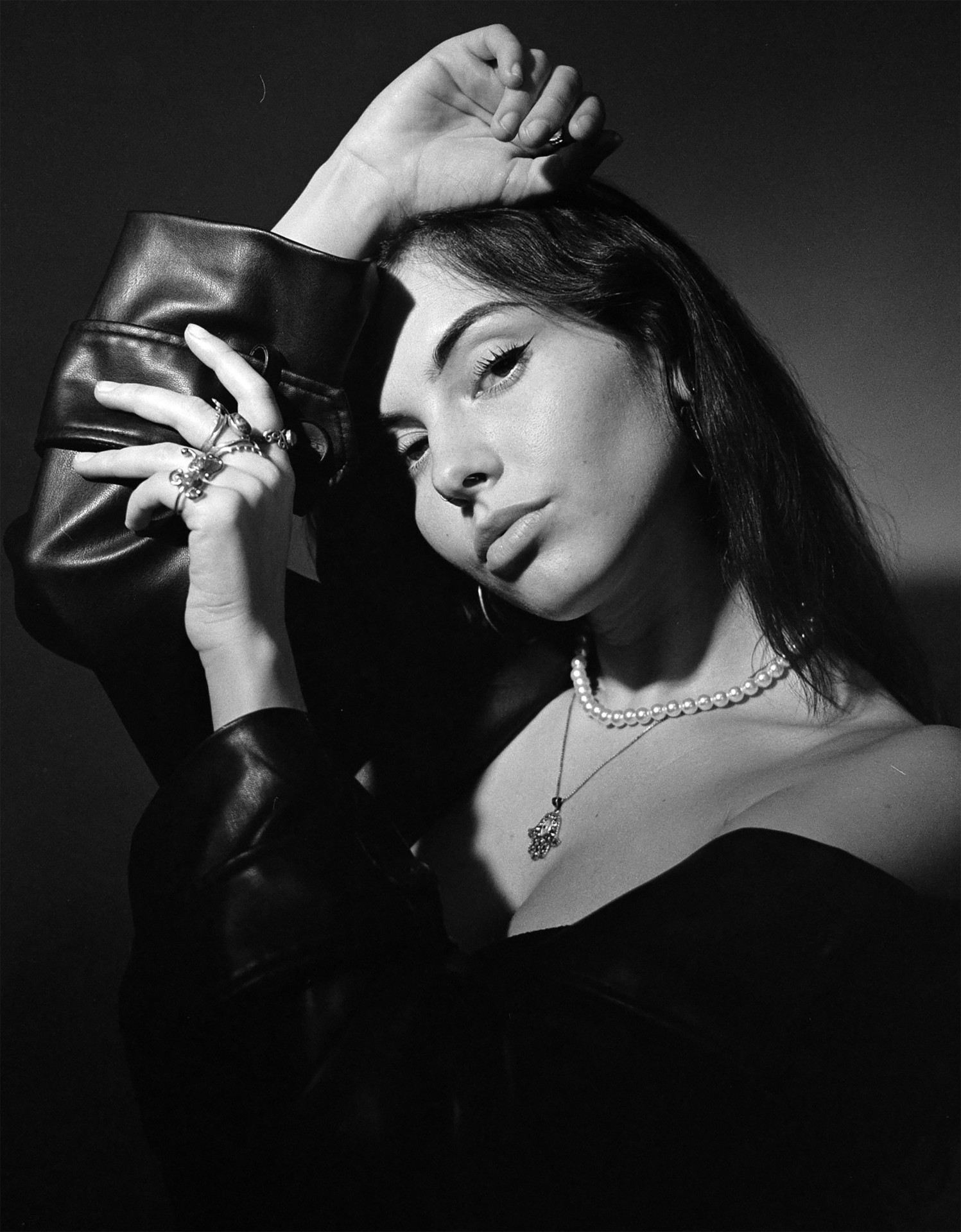 Frau mit langen Haaren in schwarzer Jacke, Hand am Kopf, trägt Perlenschmuck. Schwarz-weiß-Fotografie.