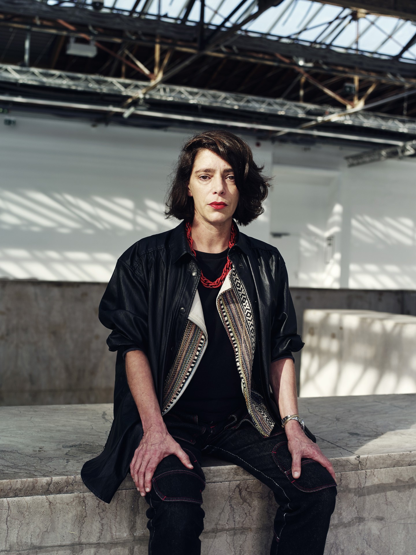 Eine Person sitzt in einem modernen Raum mit hohen Fenstern, trägt ein schwarzes Outfit und eine farbenfrohe Kette.