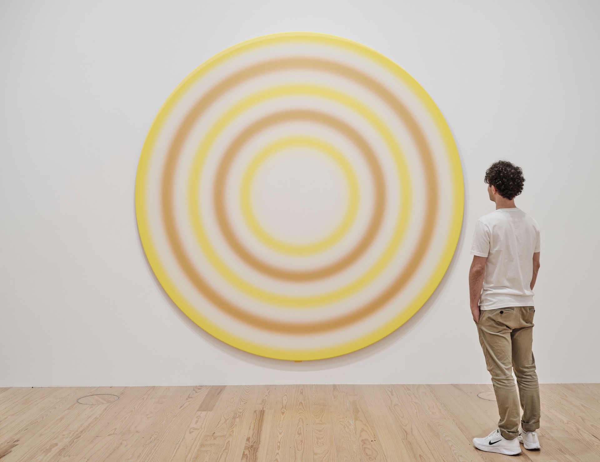 A man is looking at a round, colorful artwork with concentric circles on a white wall.