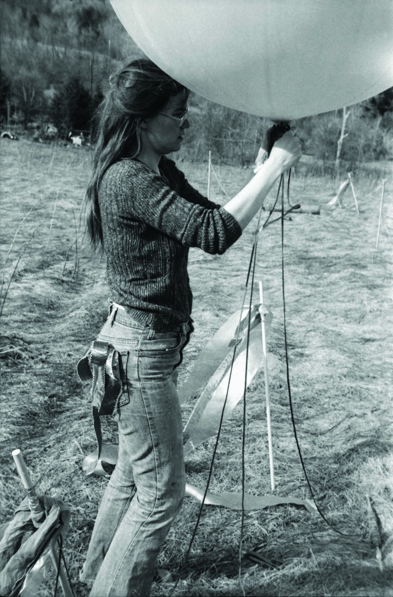 A woman with long hair and glasses ties a balloon in a field surrounded by nature and equipment.