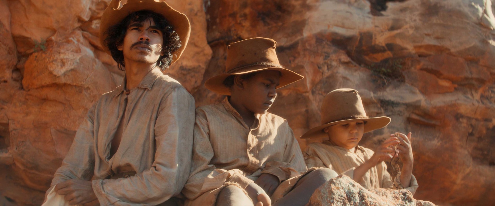 Drei Kinder und ein Mann in der Wüste sitzen auf einem Felsen, tragen Hüte und einfache Kleidung und betrachten die Umgebung.