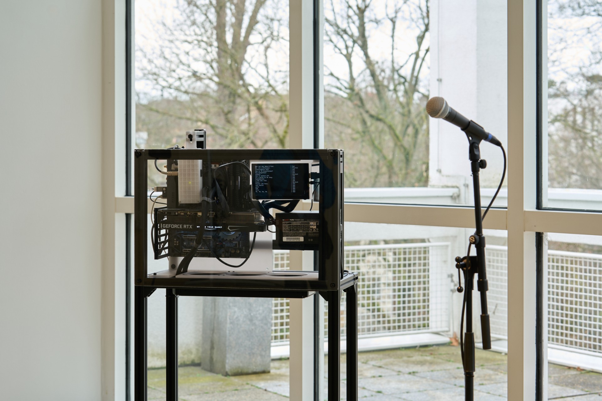 Transparenter Computer in einer Ausstellung, ausgestattet mit Mikrofon und Blick auf die Bäume draußen.