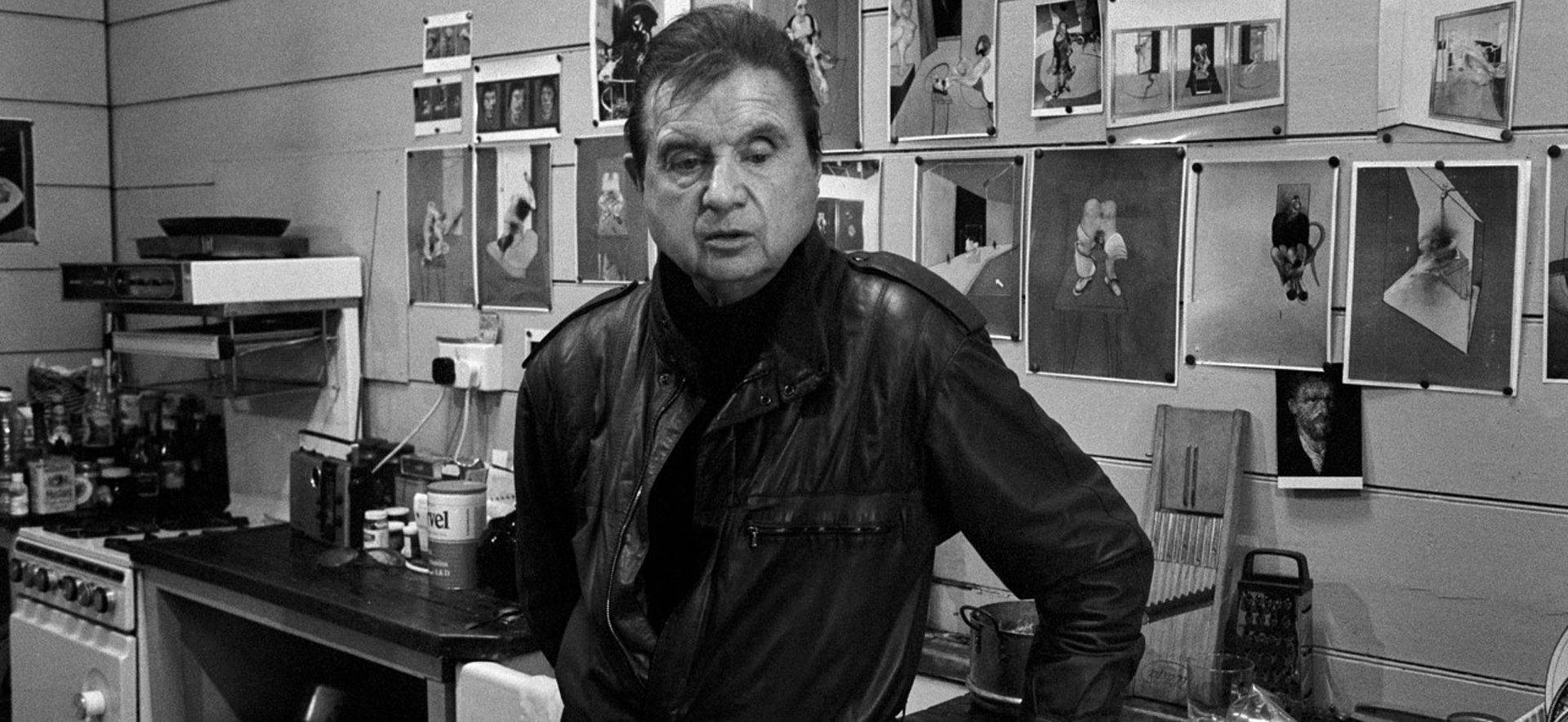 A man in a black leather jacket stands in a kitchen, surrounded by photographs on the wall and kitchen appliances.
