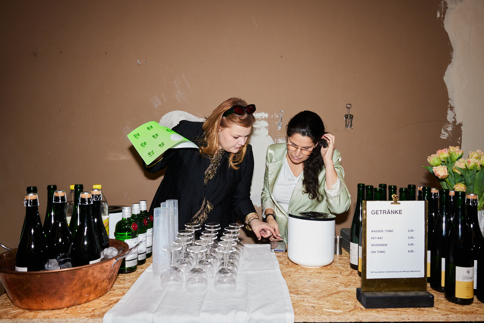 Zwei Frauen stehen an einem Getränkestand mit Flaschen, Gläsern und Preisliste. Hintergrund in neutralen Farben.
