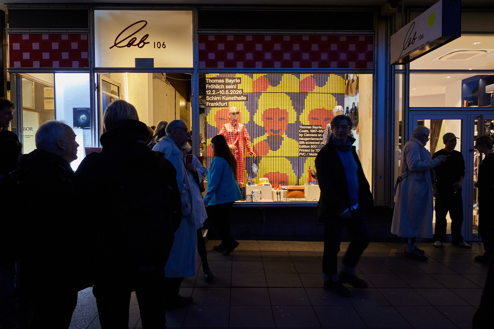 Menschen versammeln sich vor einem Kunstladen mit buntem Schaufenster und Plakaten für eine Ausstellung.