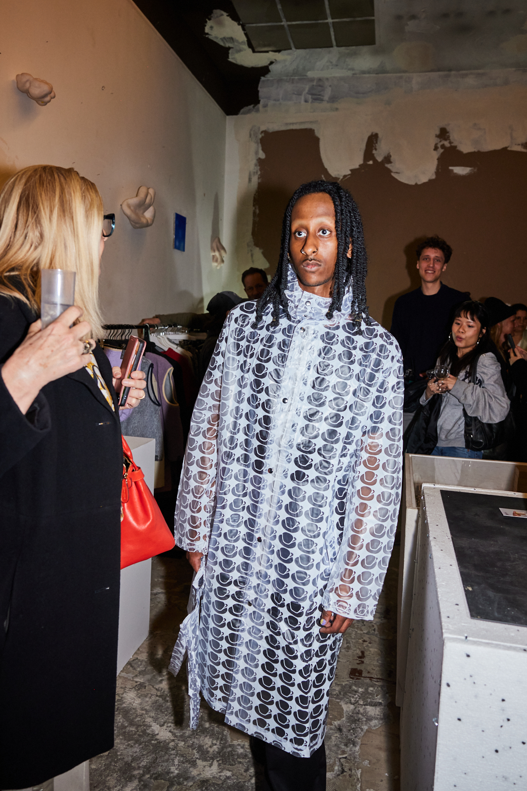 Models in einem kreativen Raum: Eine trägt einen transparenten Mantel mit Kaffeetassen-Muster, andere schauen zu.