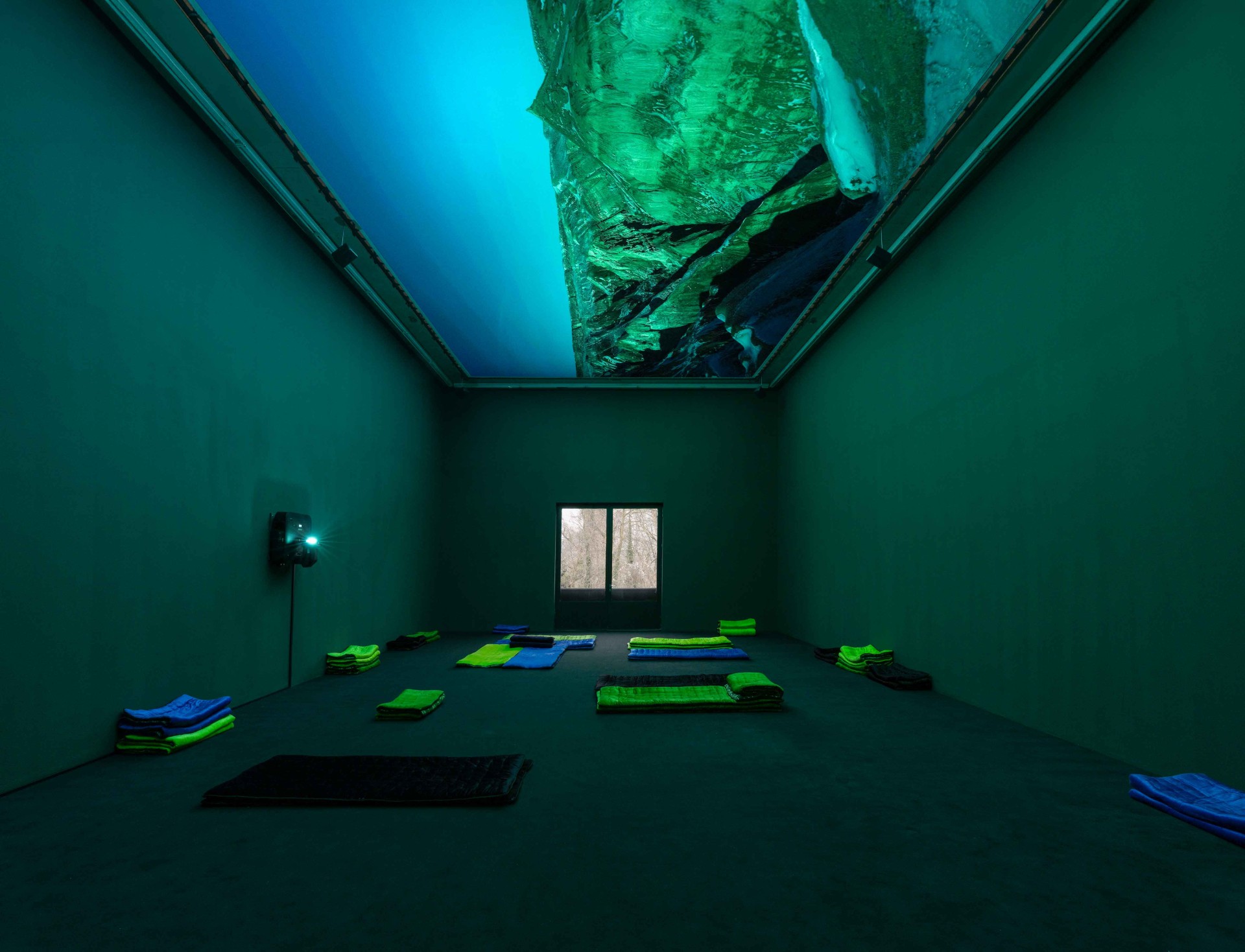 Minimalistischer Raum mit grünen Wänden, gefalteten Decken und einem blau-grünen, beleuchteten Deckengemälde.