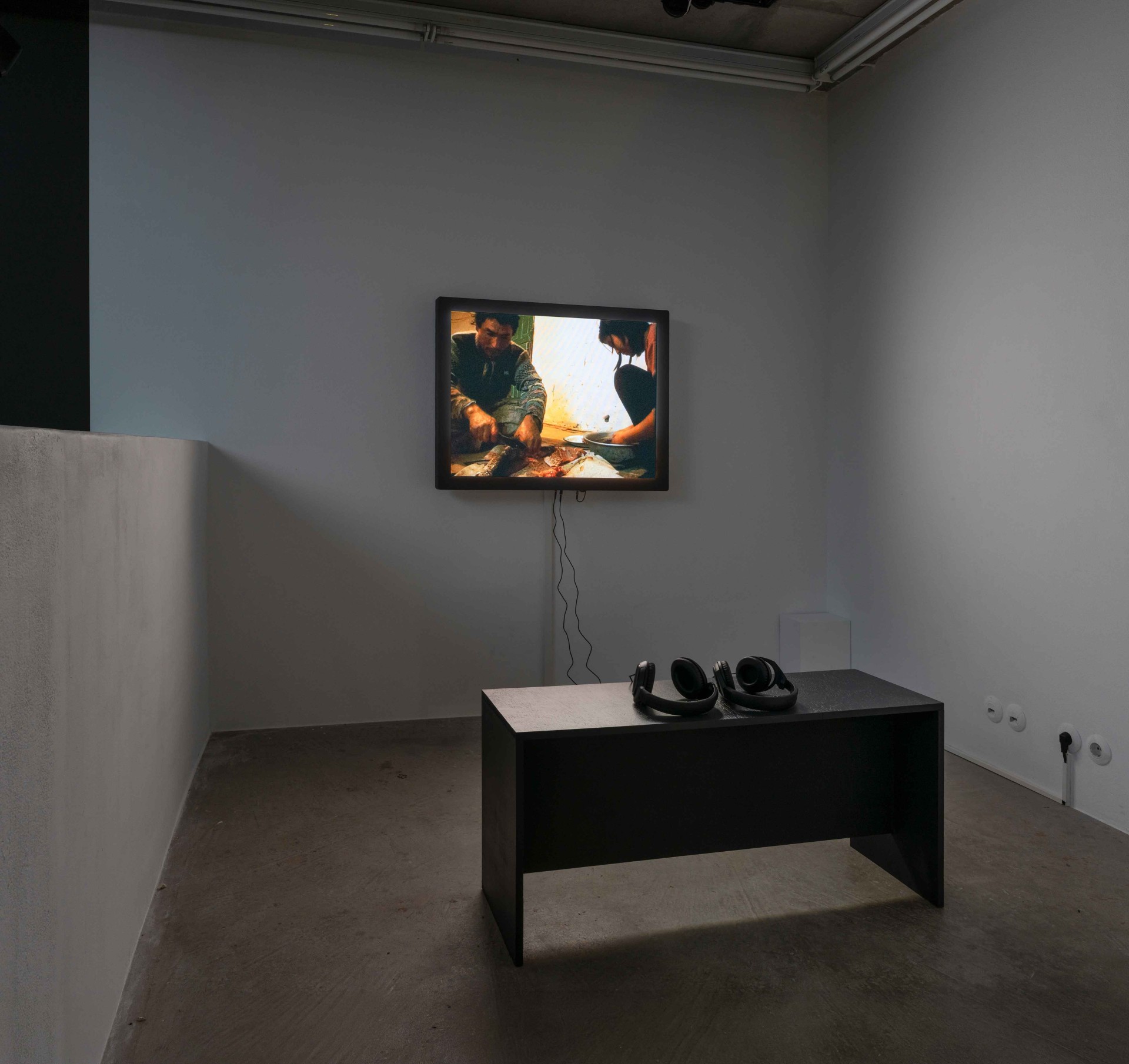 Kunstinstallation mit Fernseher, der ein Video zeigt, und einem Tisch mit Kopfhörern in einem minimalistischen Raum.