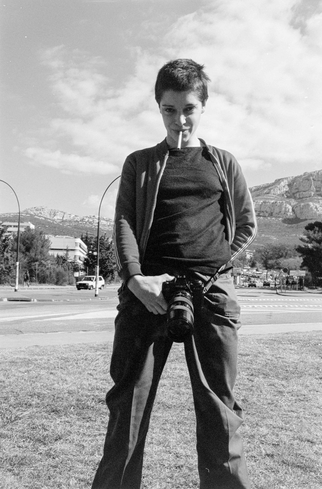 Person mit Kamera und Zigarette im Mund steht vor einer Landschaft unter blauem Himmel mit Wolken.