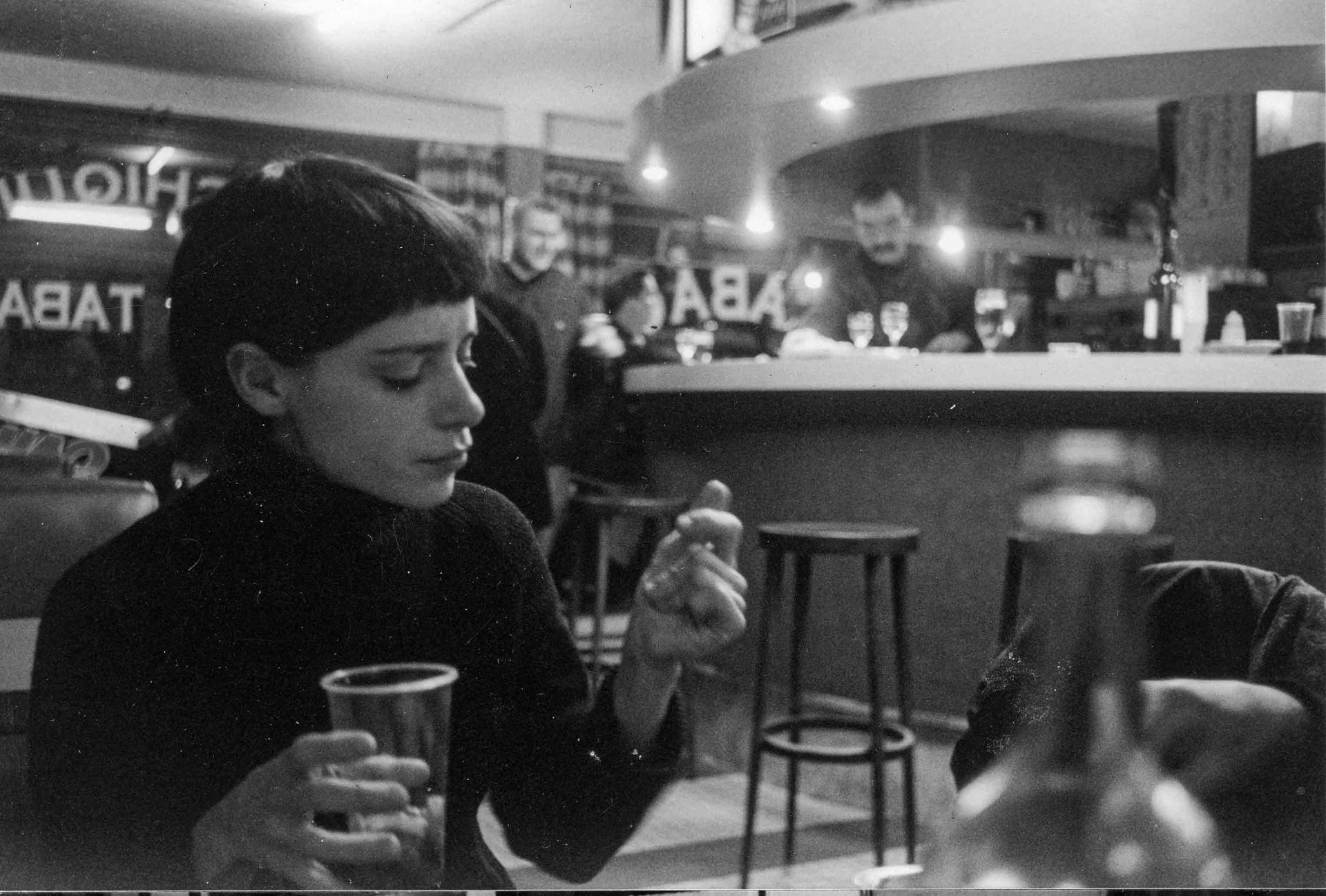 Eine nachdenkliche Person in einem Café, umgeben von Getränken und Menschen im Hintergrund. Schwarz-Weiß-Ästhetik.