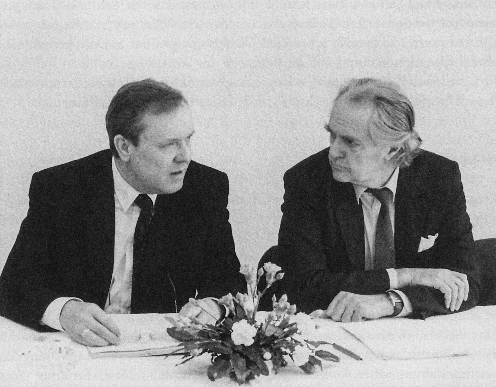 Zwei Männer in Anzügen diskutieren an einem Tisch, umgeben von Blumen und Papieren. Schwarzweißfotografie.