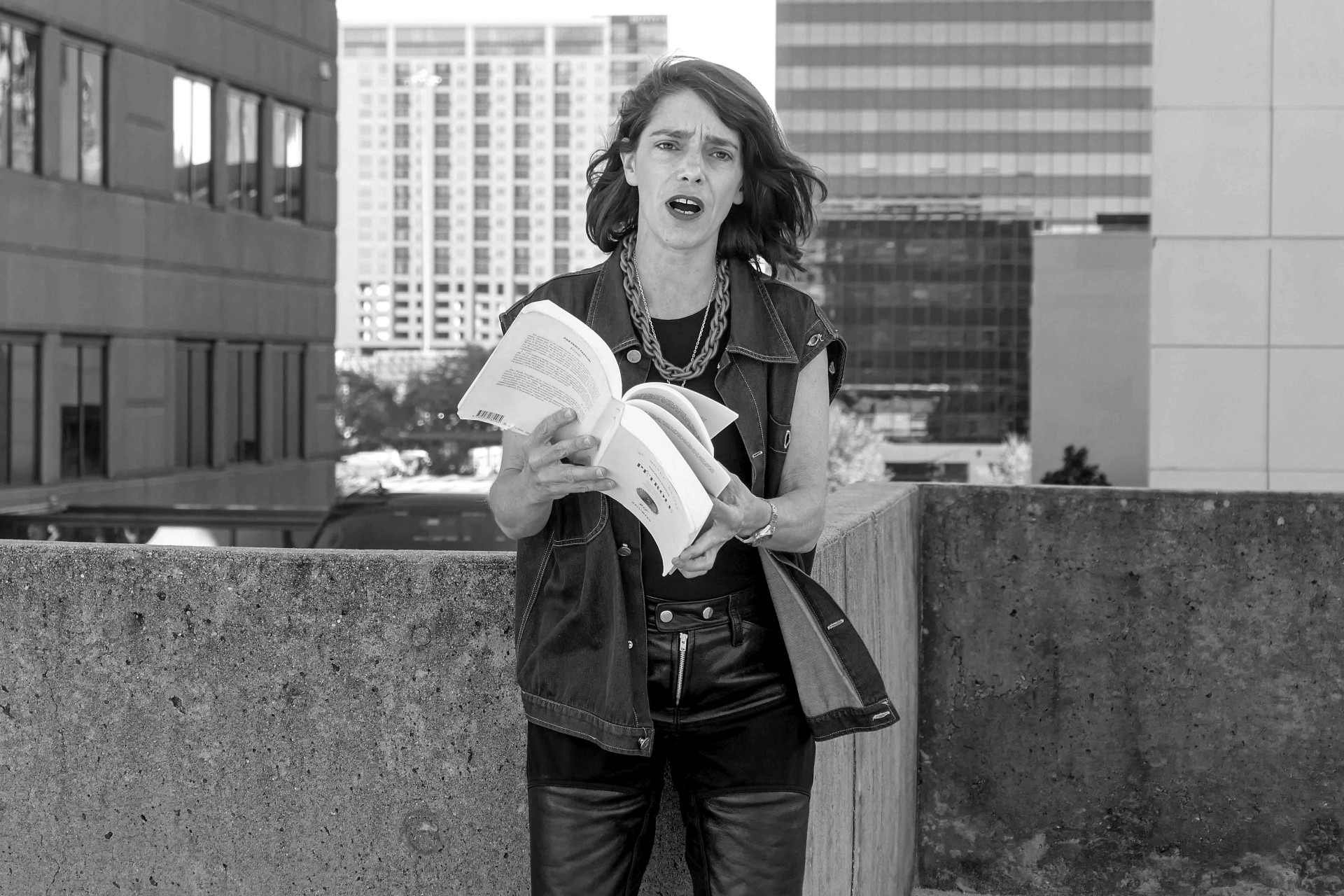 Frau in Lederhose und Jeansjacke steht auf einem Gebäude und hält ein Buch, urbane Kulisse im Hintergrund.