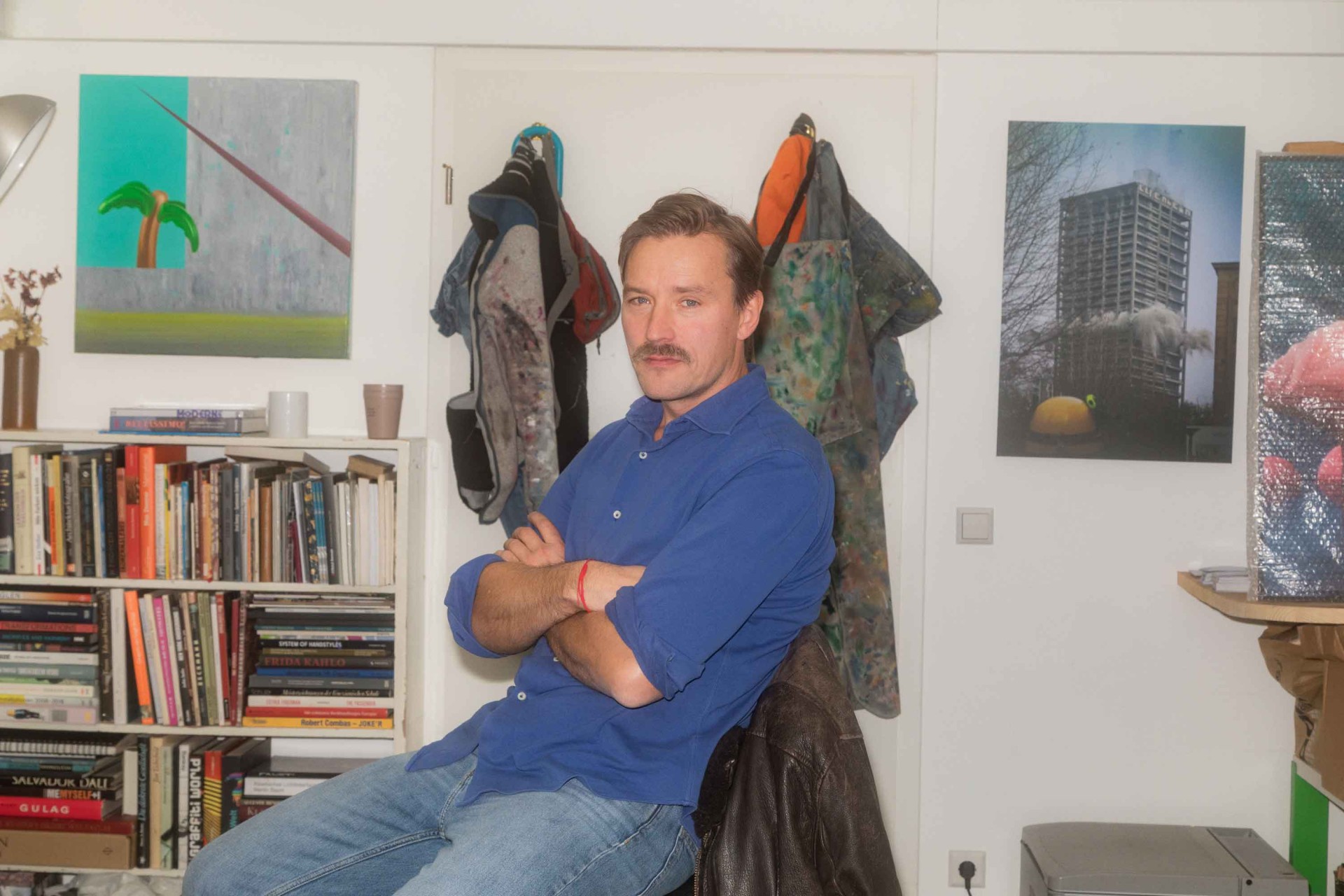 Mann mit Schnurrbart sitzt nachdenklich im Atelier, umgeben von Kunst und Büchern.