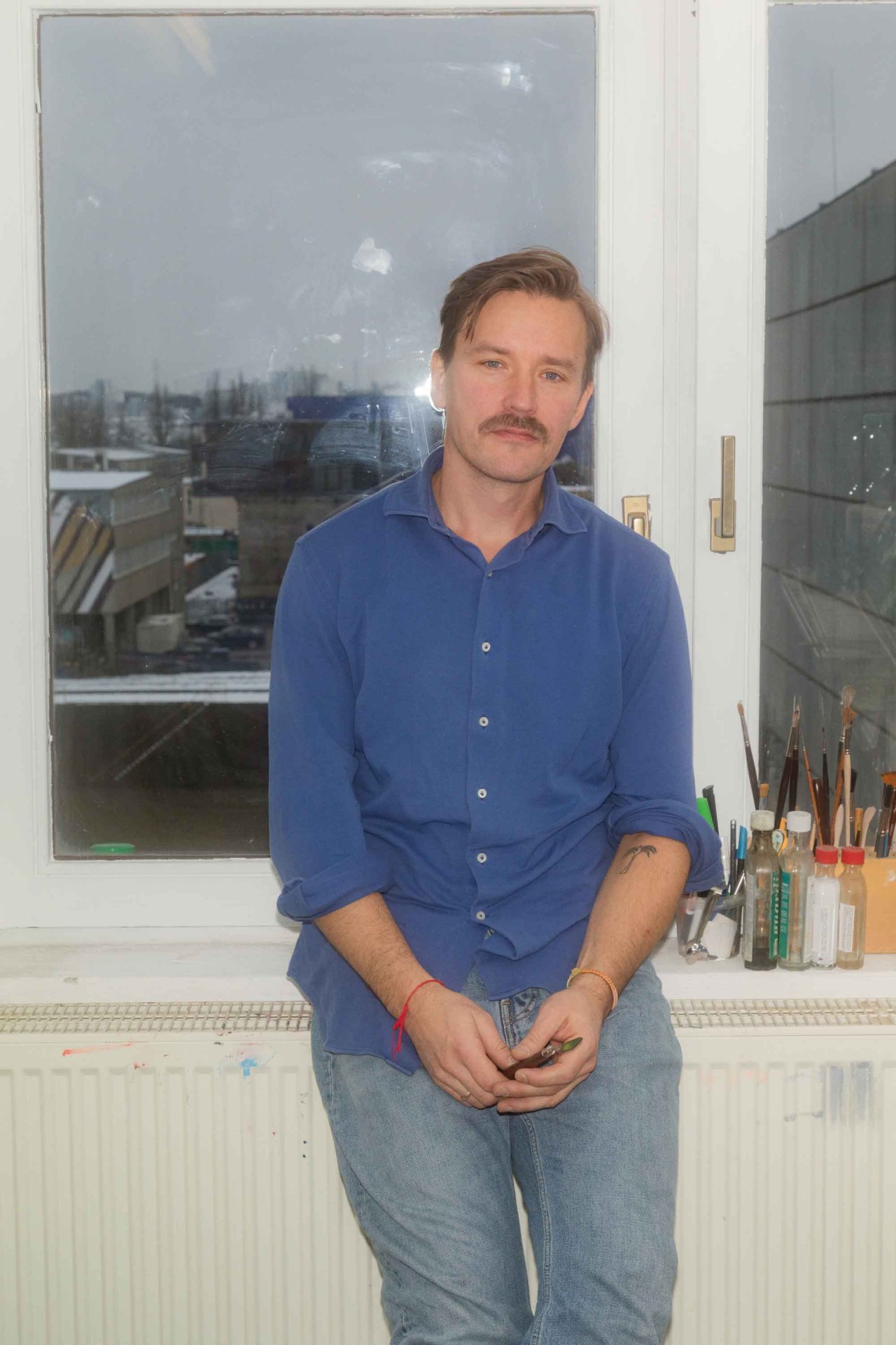 Mann mit Schnurrbart in blauem Hemd und Jeans sitzt am Fenster mit Blick auf die Stadt und Malutensilien.