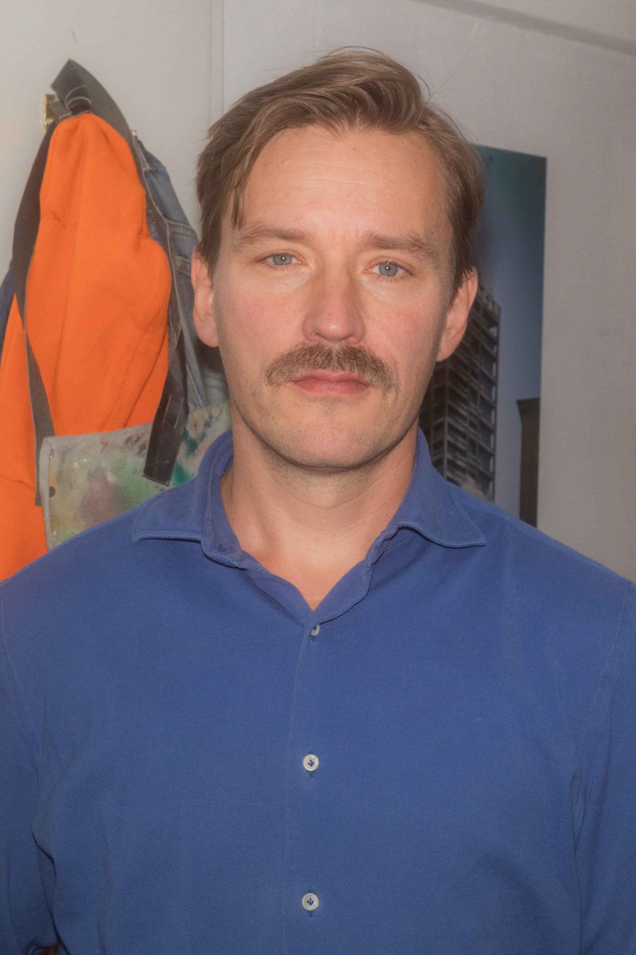 Ein Mann mit blauen Augen und Schnurrbart trägt ein blaues Hemd, im Hintergrund sind eine orange Jacke und ein Bild.