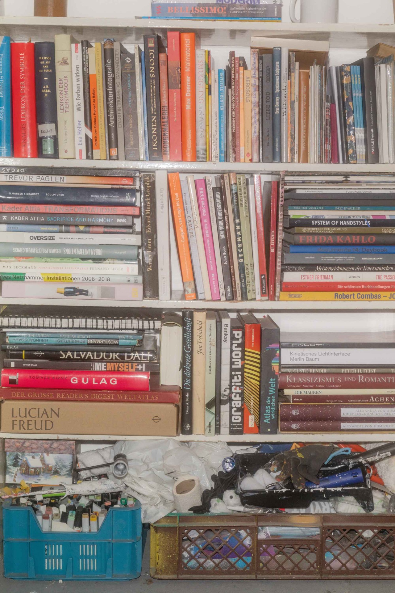 Ein Regal mit vielen Büchern und Kunstwerken, daneben Kisten mit Materialien und persönlichen Gegenständen.