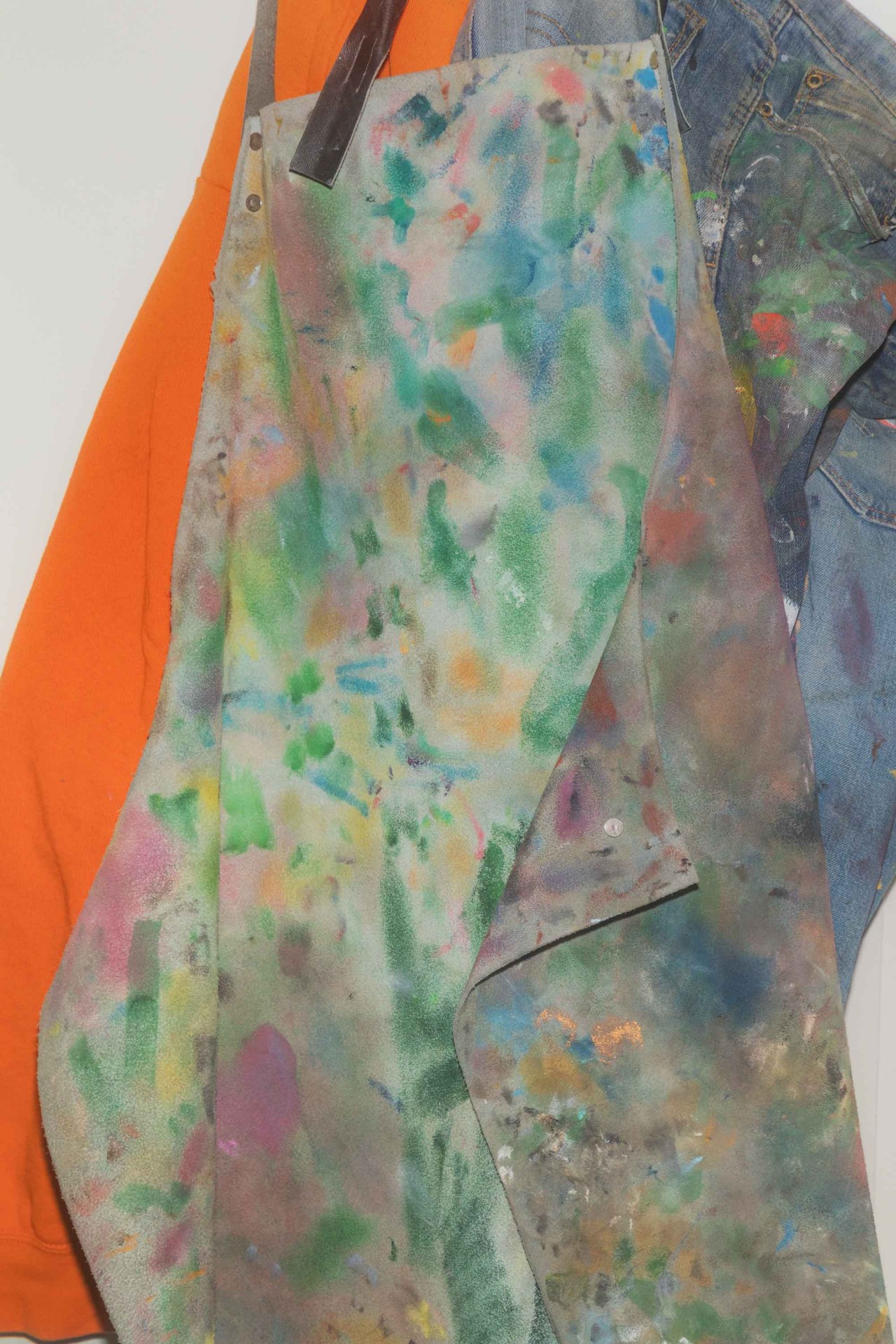 Bunt besprenkelte Schürze hängt an einer Wand, neben einer orangefarbenen Jacke und einer blauen Jeansjacke.