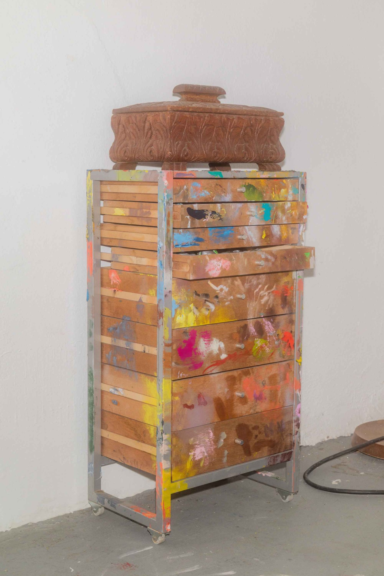 Ein Holztisch mit bunten Farbspritzern und einer dekorativen Schale darauf, vor einer weißen Wand.