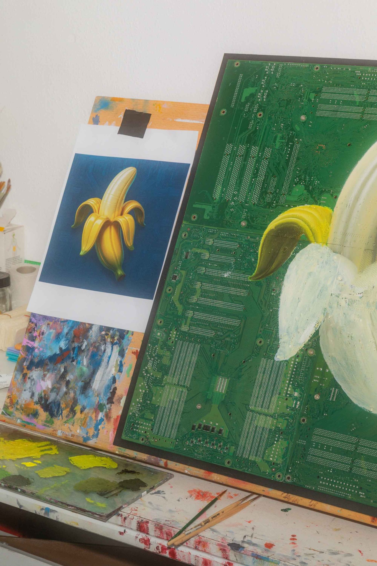 Kunstwerk mit einer Banane auf einer grünen Leiterplatte, umgeben von Farbpaletten und Malutensilien.