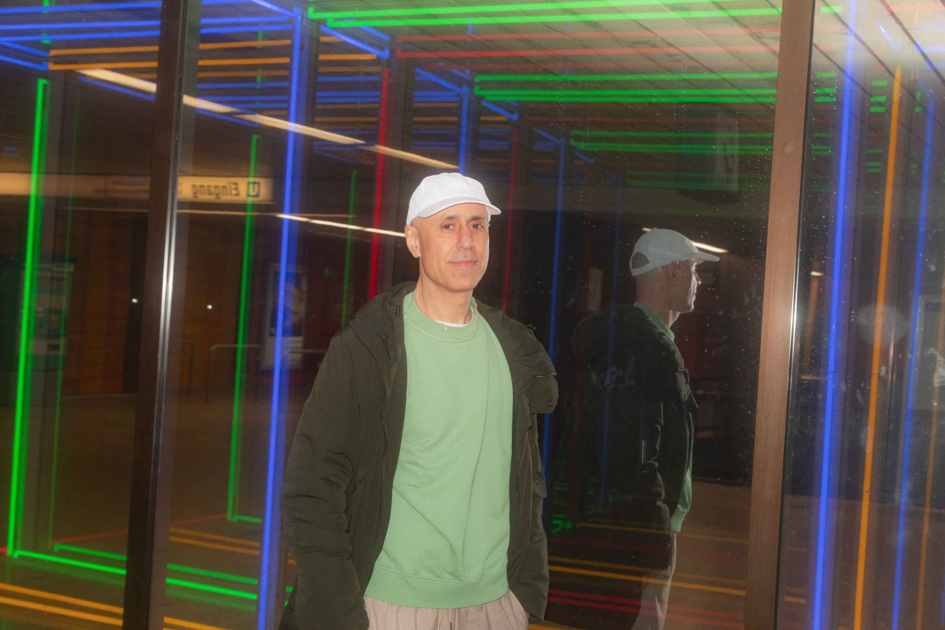Mann in weißer Kappe und grünem Sweatshirt steht vor bunten Neonlichtern und einem Spiegel.