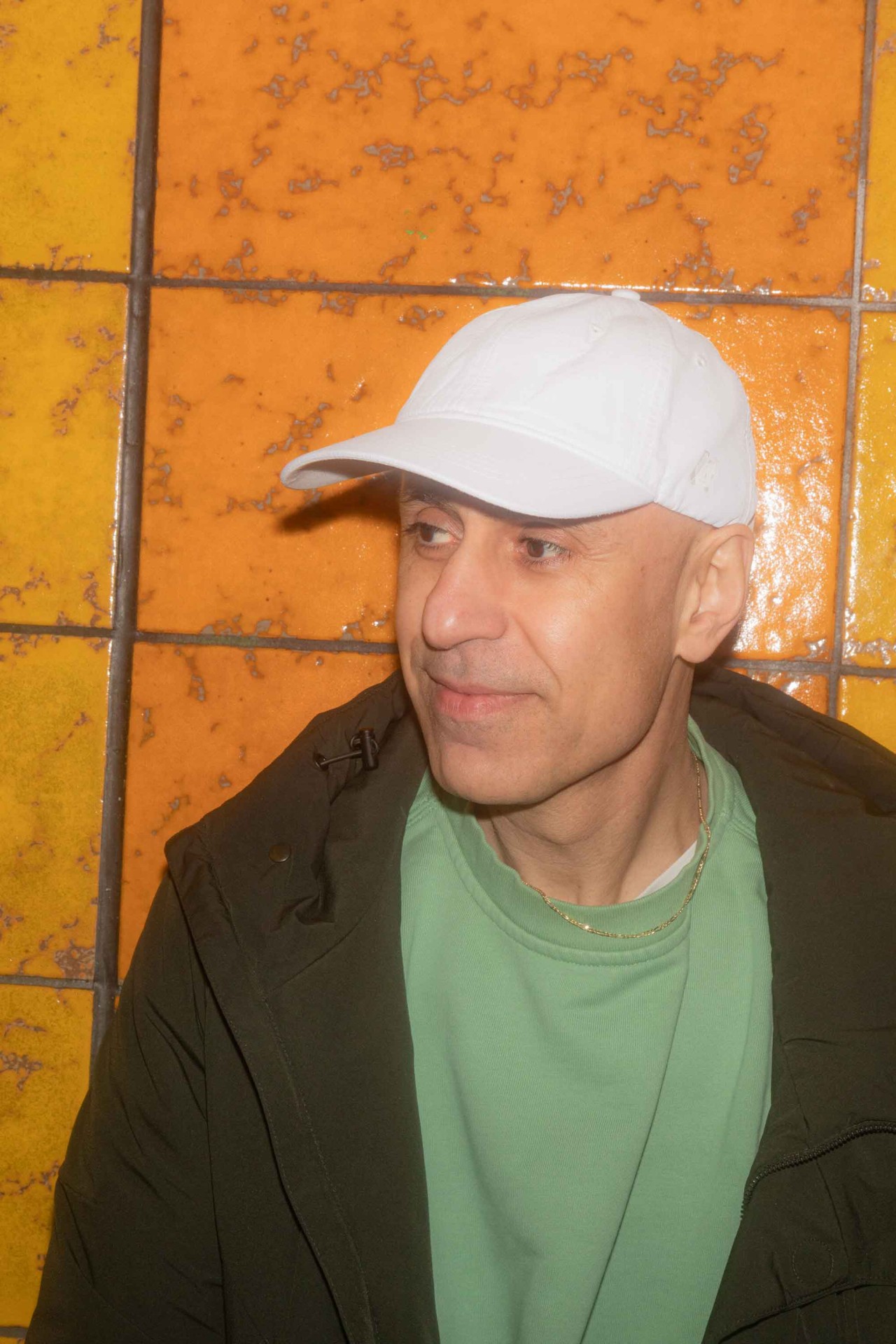 Mann mit weißer Mütze und grünem Pullover vor einer bunten Wand aus gelben und orangefarbenen Fliesen.