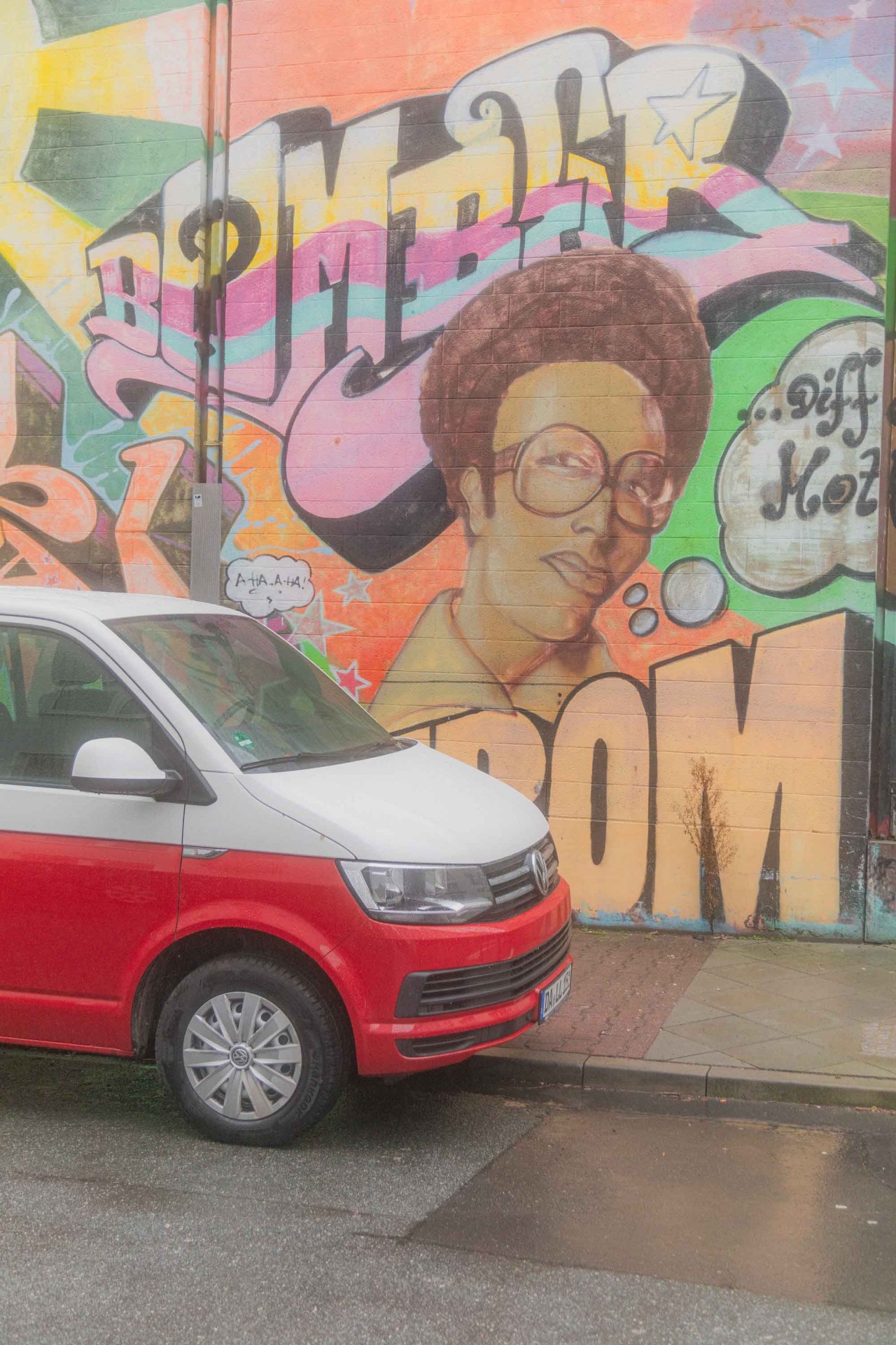 Rotes und weißes Auto vor einer bunten Wand mit Graffiti, Porträt und Schriftzug.
