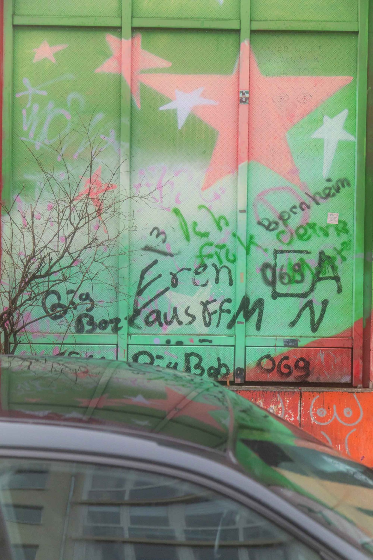 Graffiti auf einer grünen Wand mit bunten Sternen, darunter Sträucher und ein Teil eines Autos.