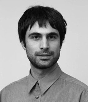 Porträt eines Mannes mit mittellangen Haaren, der in die Kamera blickt, vor einem neutralen Hintergrund. Schwarz-weiß.