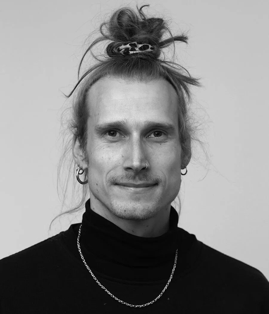 Porträt eines Mannes mit Dutt, Piercing und Halskette vor neutralem Hintergrund.