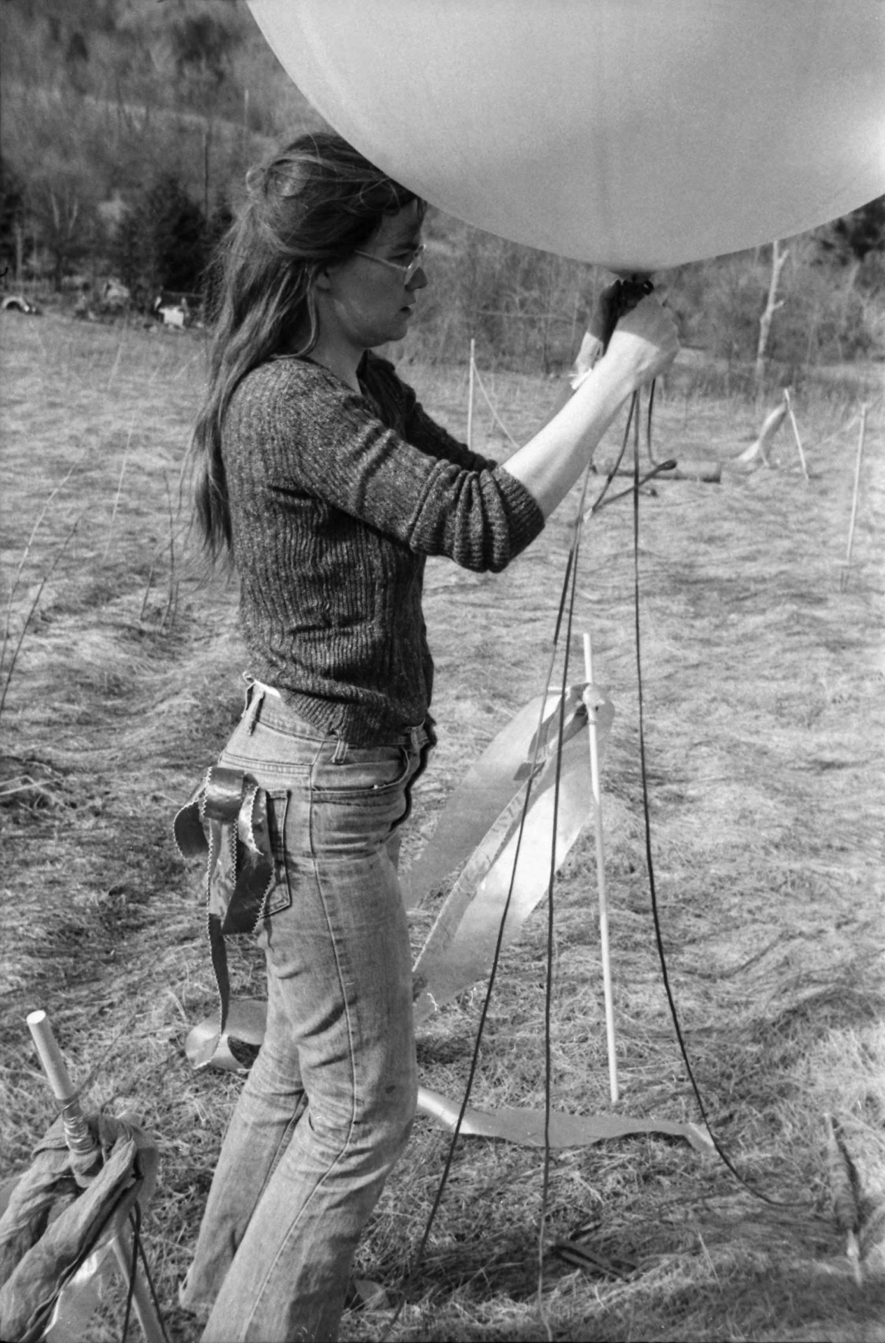 Frau mit langen Haaren und Brille bereitet einen großen Ballon auf einem Feld vor. Schwarz-Weiß-Foto.
