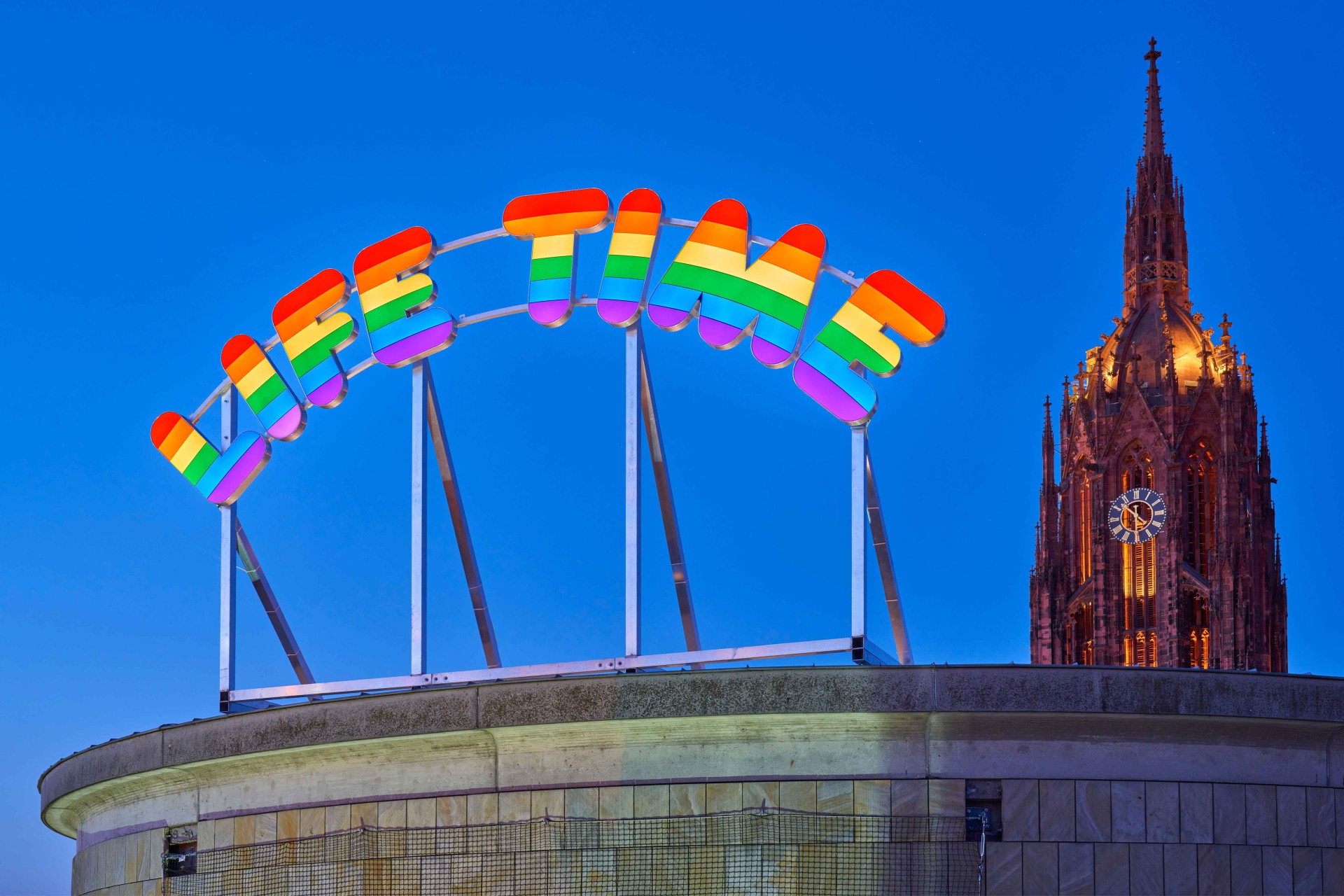 Bunt leuchtendes "LIFE TIME"-Schild vor blauem Himmel, im Hintergrund der beleuchtete Kirchturm.