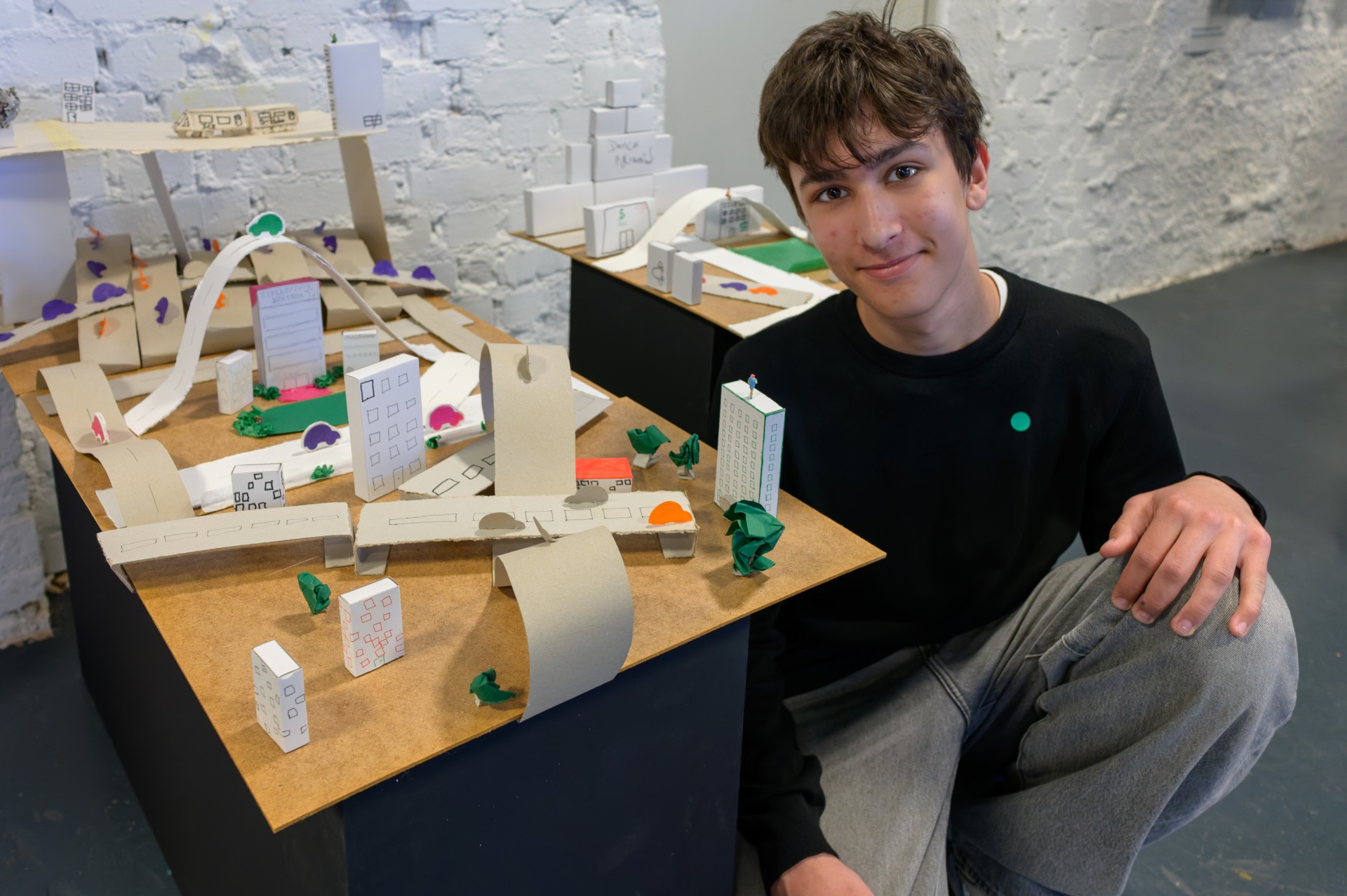 Junger Mann neben einem kreativen Stadtmodell aus Papier und Knete in einem Ausstellungsraum.