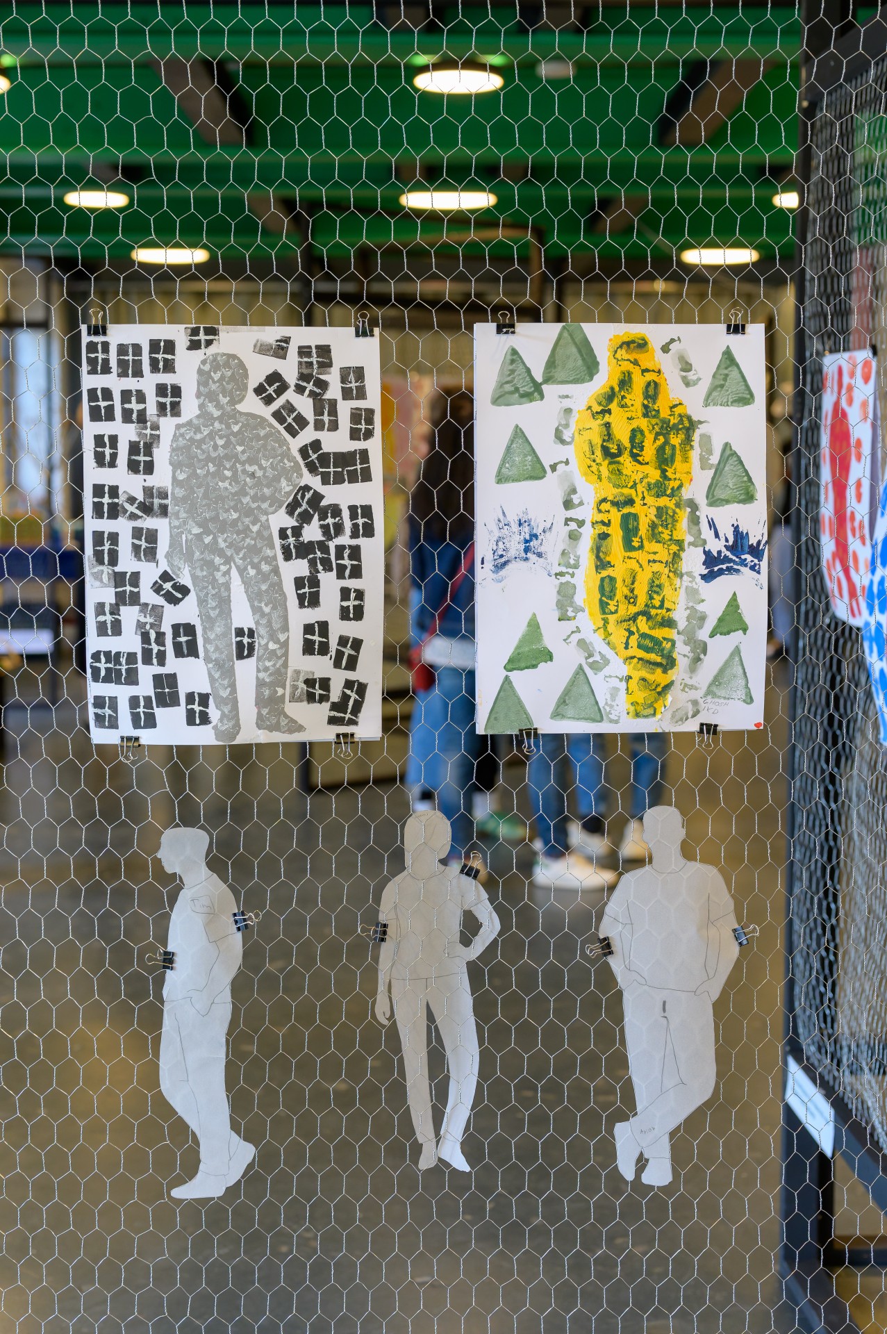 Zwei Papierkunstwerke mit Silhouetten und geometrischen Formen hinter einem Maschendrahtzaun in einer Galerie.