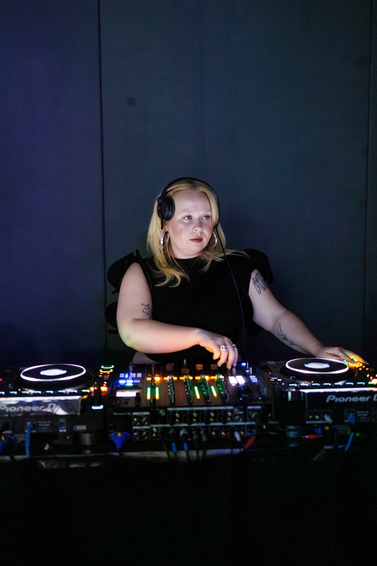DJ in a black outfit mixing music on a mixer, surrounded by colorful lights, with a focused gaze on the turntables.