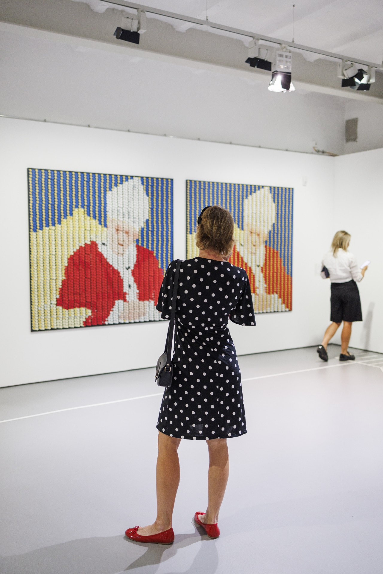 Frau im gepunkteten Kleid betrachtet Kunst in einer Galerie. Im Hintergrund eine weitere Besucherin mit Smartphone.