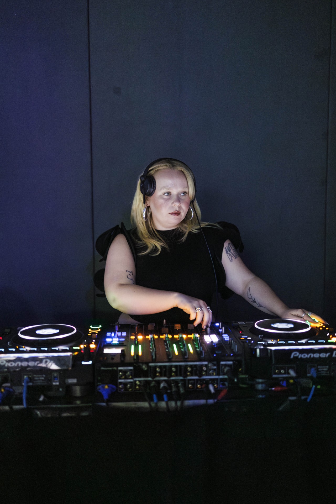 DJ mit Kopfhörern am Mischpult vor dunklem Hintergrund mit bunten Lichtern.