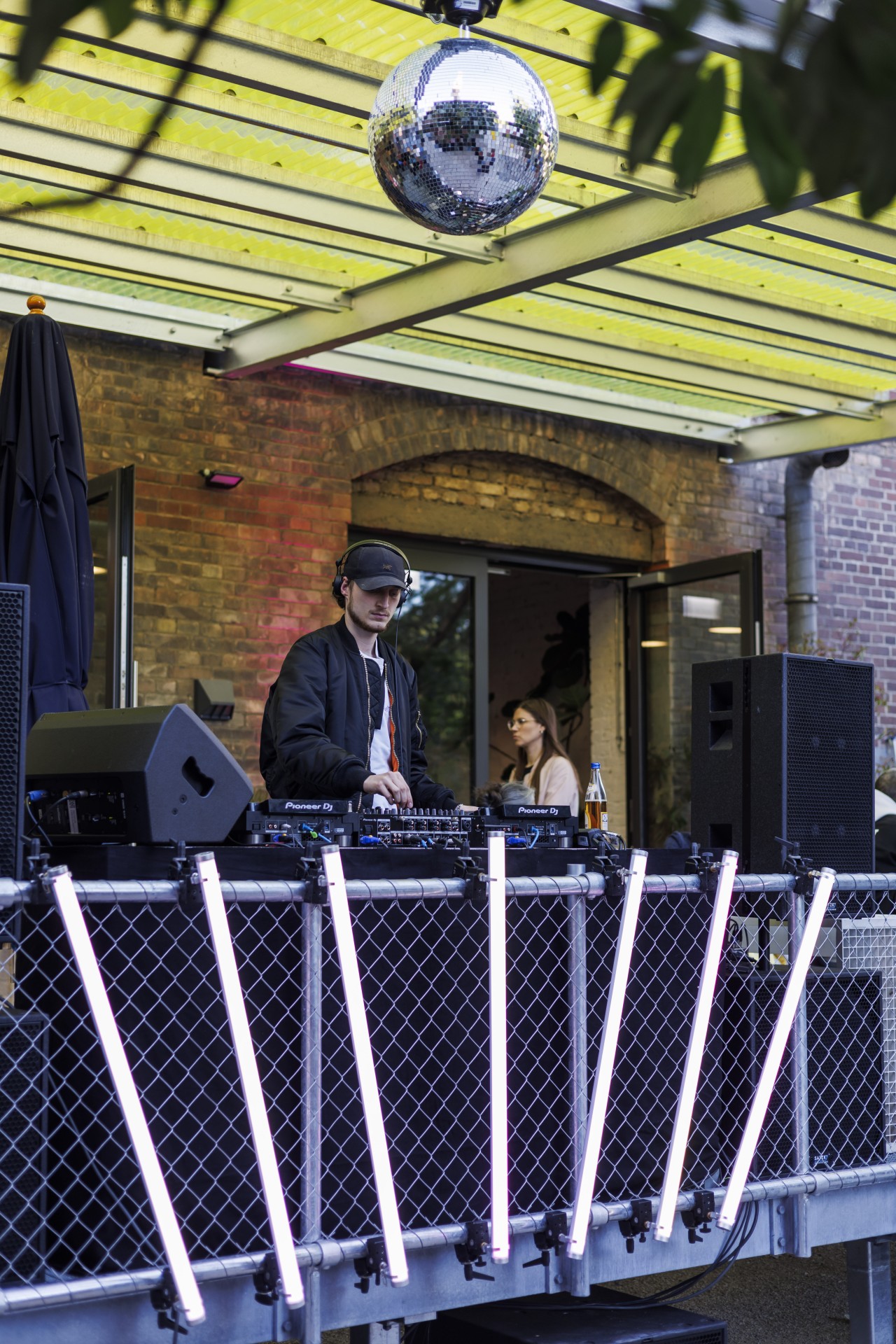 DJ spielt an einem Pult unter einem Glasdach, im Hintergrund ein Discoball und Neonbeleuchtung.